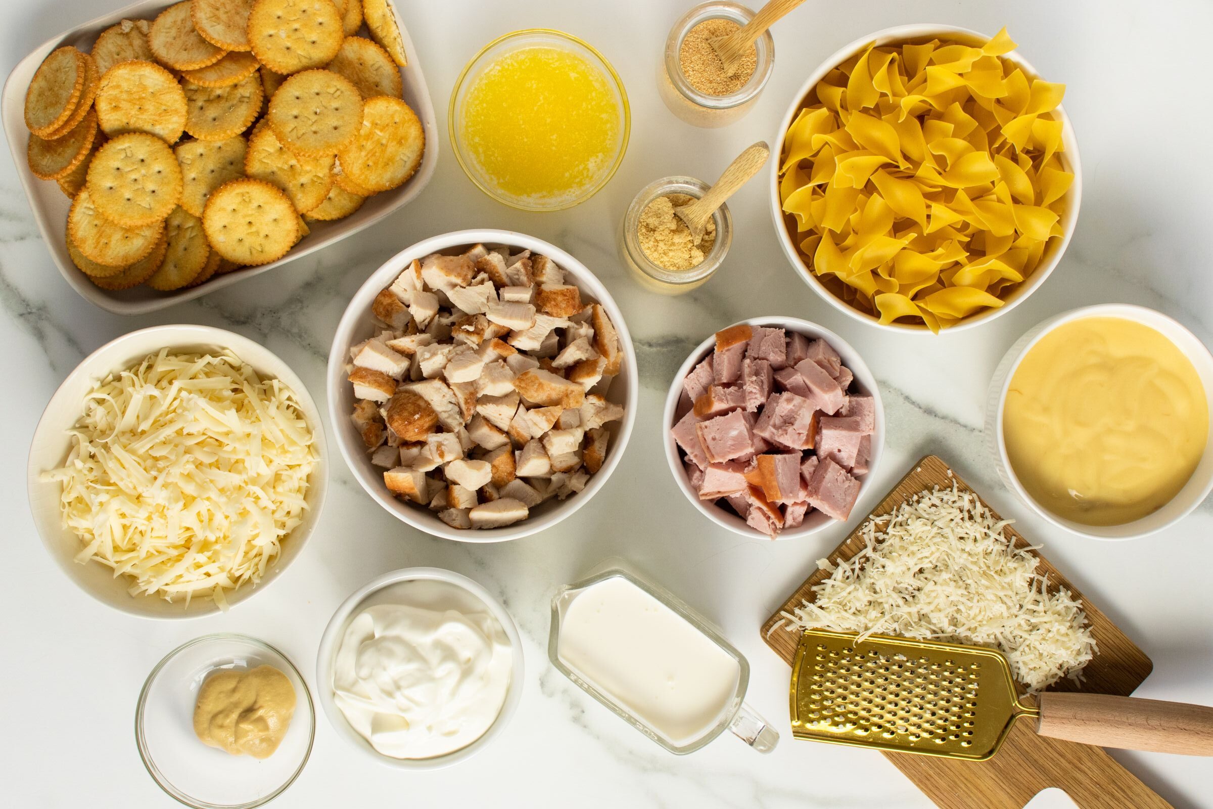 An overhead view of neatly arranged ingredients for a casserole, including uncooked egg noodles, shredded cheese, diced ham, diced chicken, crackers, melted butter, milk, cream soup, sour cream, mustard, and seasonings in bowls.