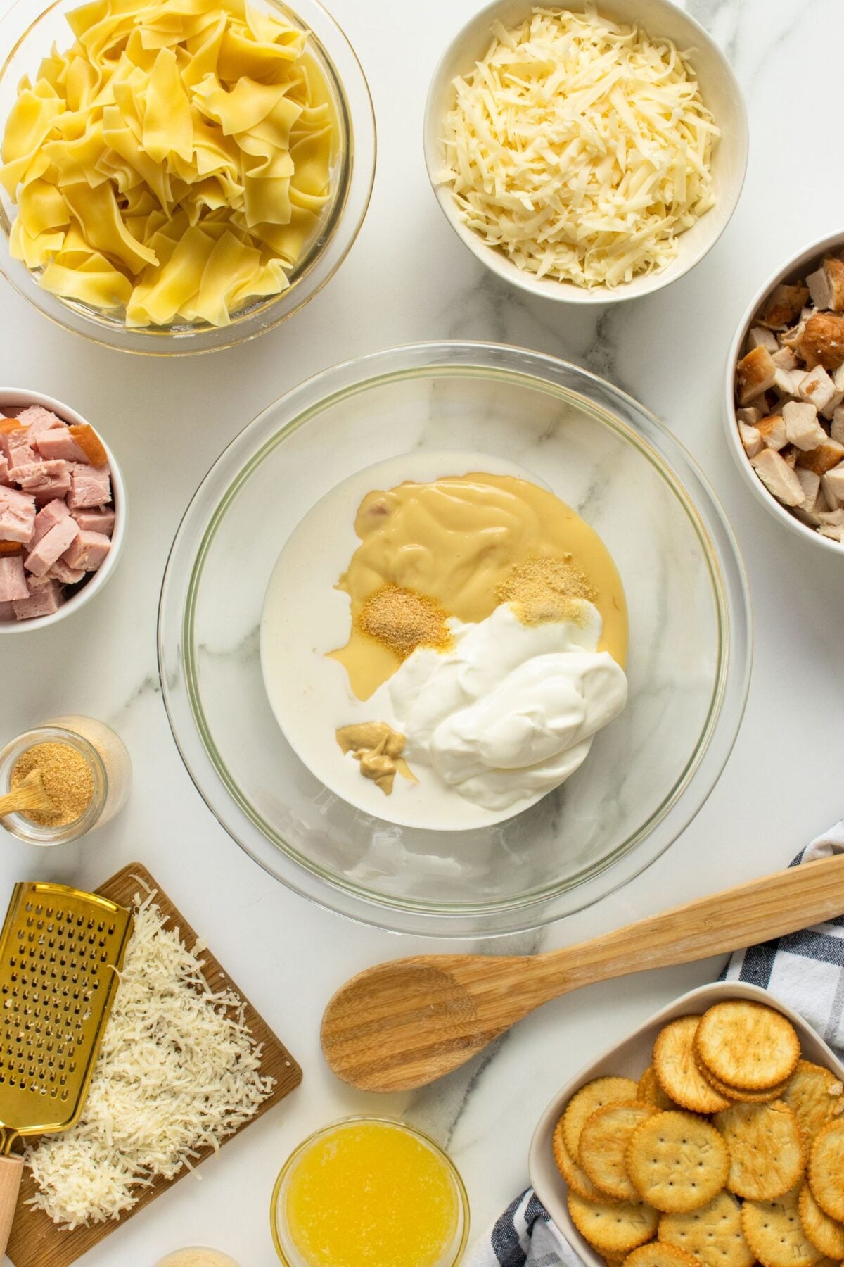 Bowls with ingredients for a casserole on a white surface: uncooked egg noodles, shredded cheese, diced ham and chicken, creamy sauce in a large bowl, grated cheese, wooden spoon, butter, and round crackers.