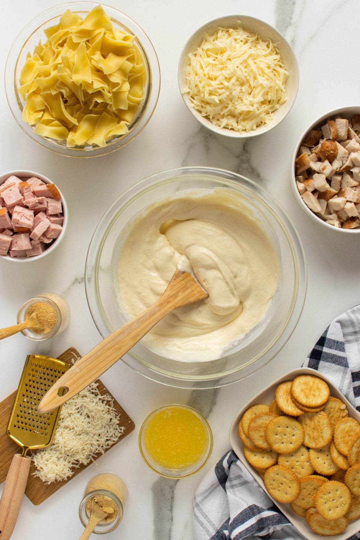 A bowl of creamy sauce being mixed with a spatula is surrounded by bowls of pasta, shredded cheese, cubed ham, cubed chicken, crackers, melted butter, seasonings, and a cheese grater on a marble surface.