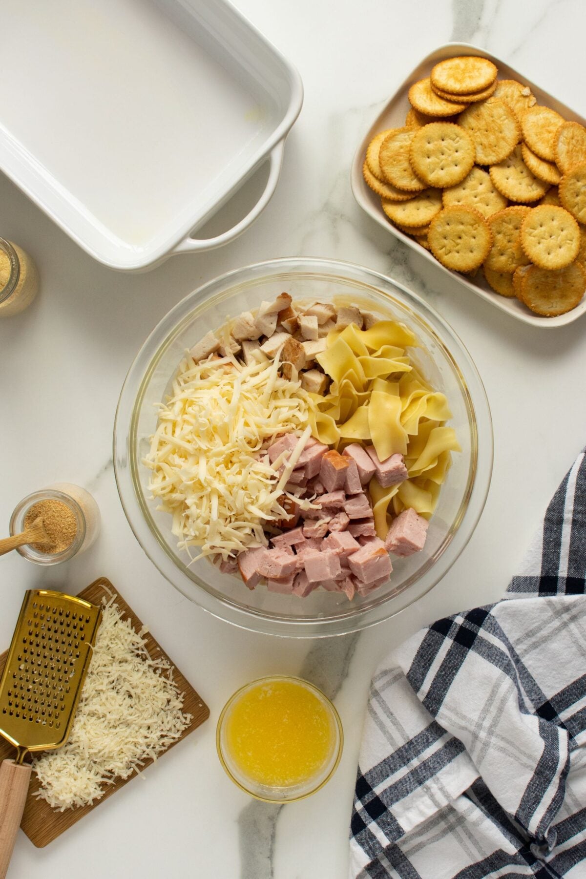 A bowl with shredded cheese, cubed ham, and uncooked noodles sits on a countertop near a dish of Ritz crackers, a small bowl of melted butter, a pile of grated cheese, and a striped kitchen towel.