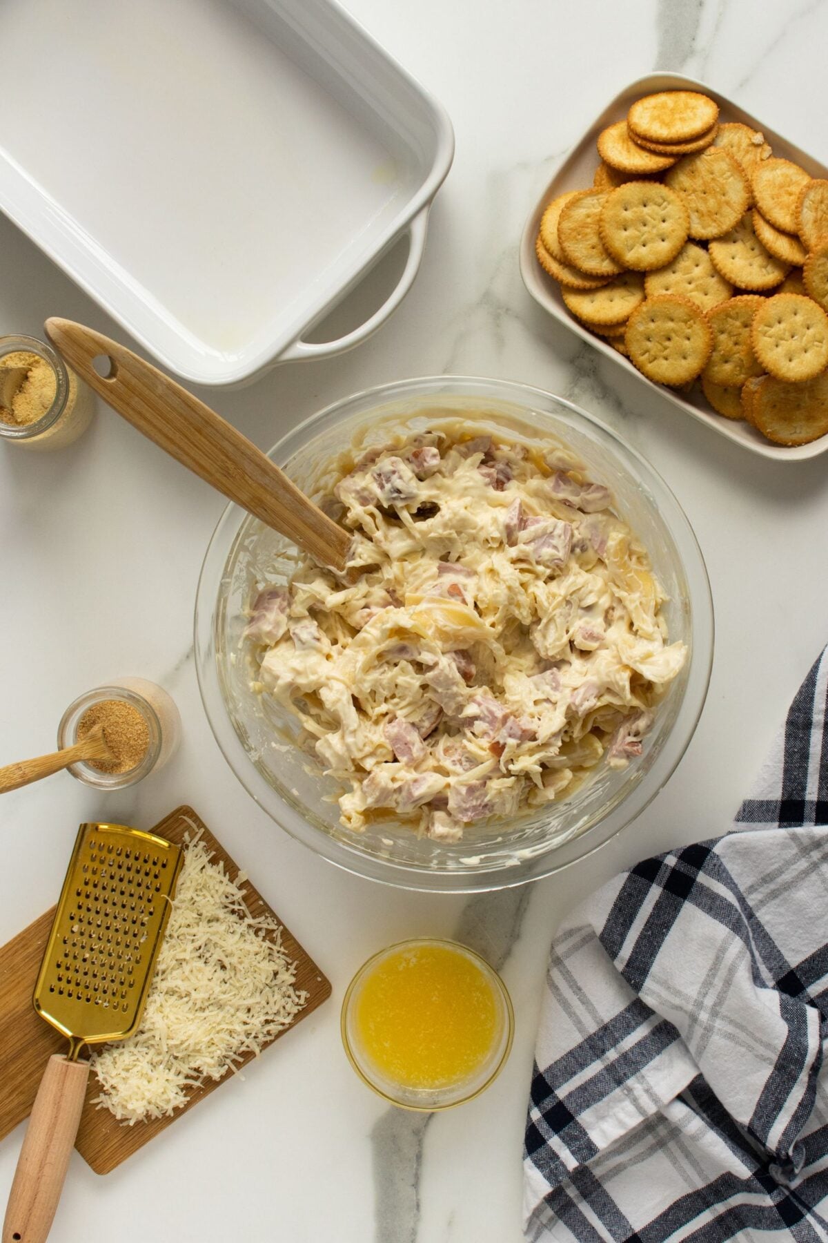 A glass bowl filled with a creamy mixture and a wooden spoon sits on a marble countertop, surrounded by grated cheese, melted butter, seasonings, a dish of round crackers, and a baking dish. A checkered towel is nearby.