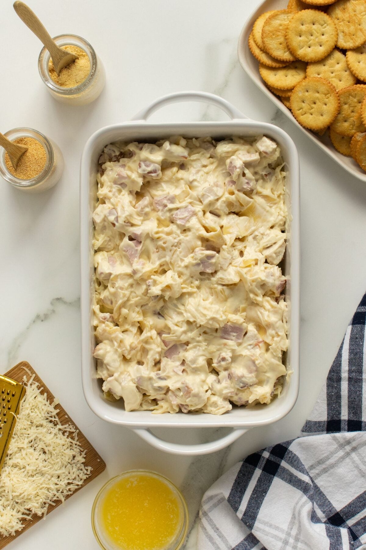 A white casserole dish filled with a creamy noodle and ham mixture is on a marble surface. Surrounding it are jars of seasoning, a plate of round crackers, shredded cheese, melted butter, and a striped cloth.
