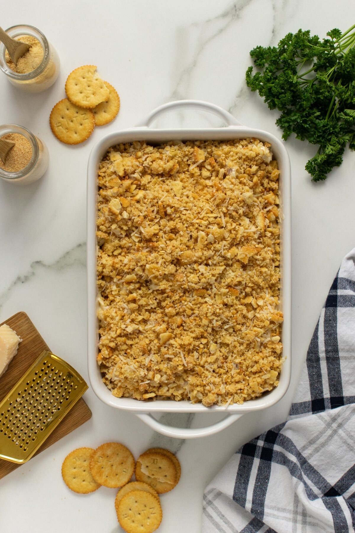 A white baking dish filled with a casserole topped with crushed crackers sits on a marble surface. Surrounding it are whole crackers, a cheese grater, parsley, a jar of seasoning, and a striped kitchen towel.