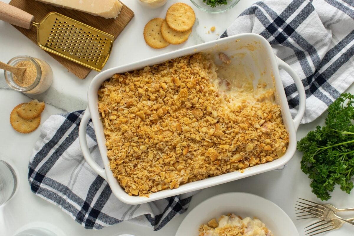 A baking dish with a creamy casserole topped with crushed crackers, one serving missing. Surrounding the dish are crackers, grated cheese, parsley, a striped towel, and kitchen utensils on a white surface.