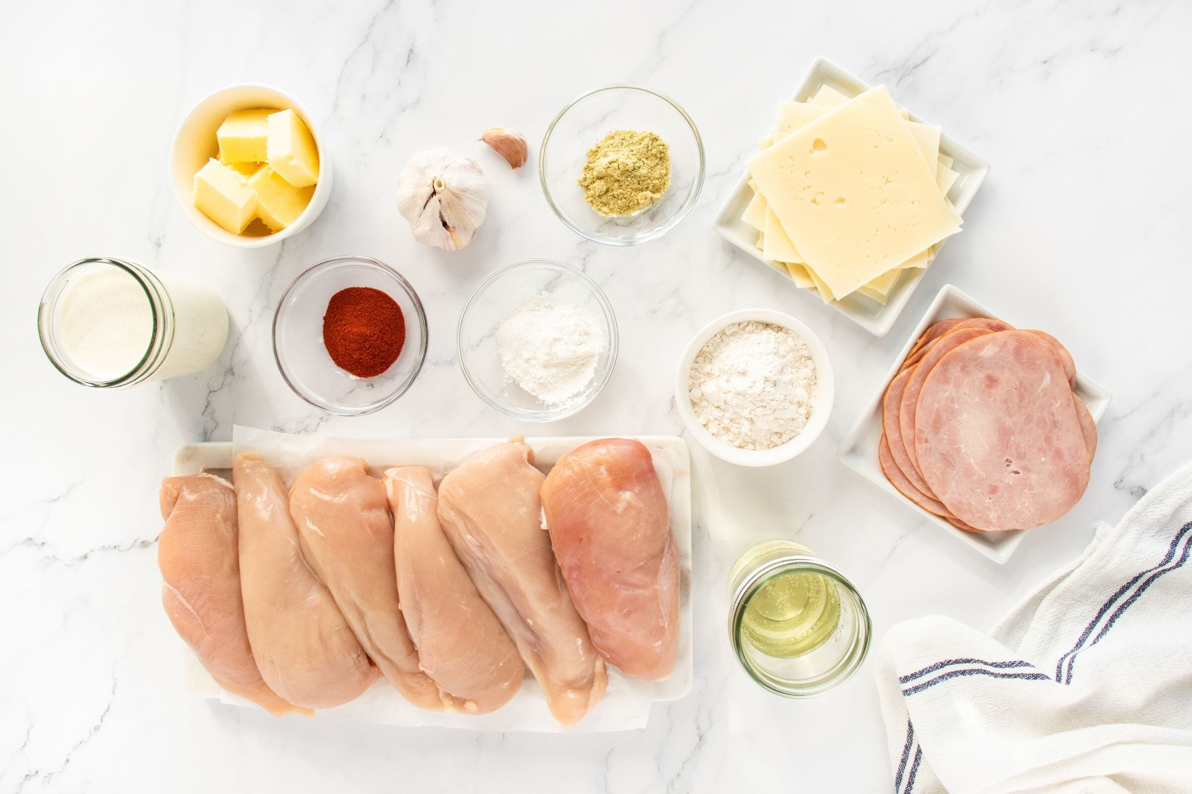 Raw chicken breasts, sliced ham, Swiss cheese, butter, garlic, flour, oil, cream, paprika, ground seasoning, and cornstarch are neatly arranged on a white surface with a striped kitchen towel nearby.