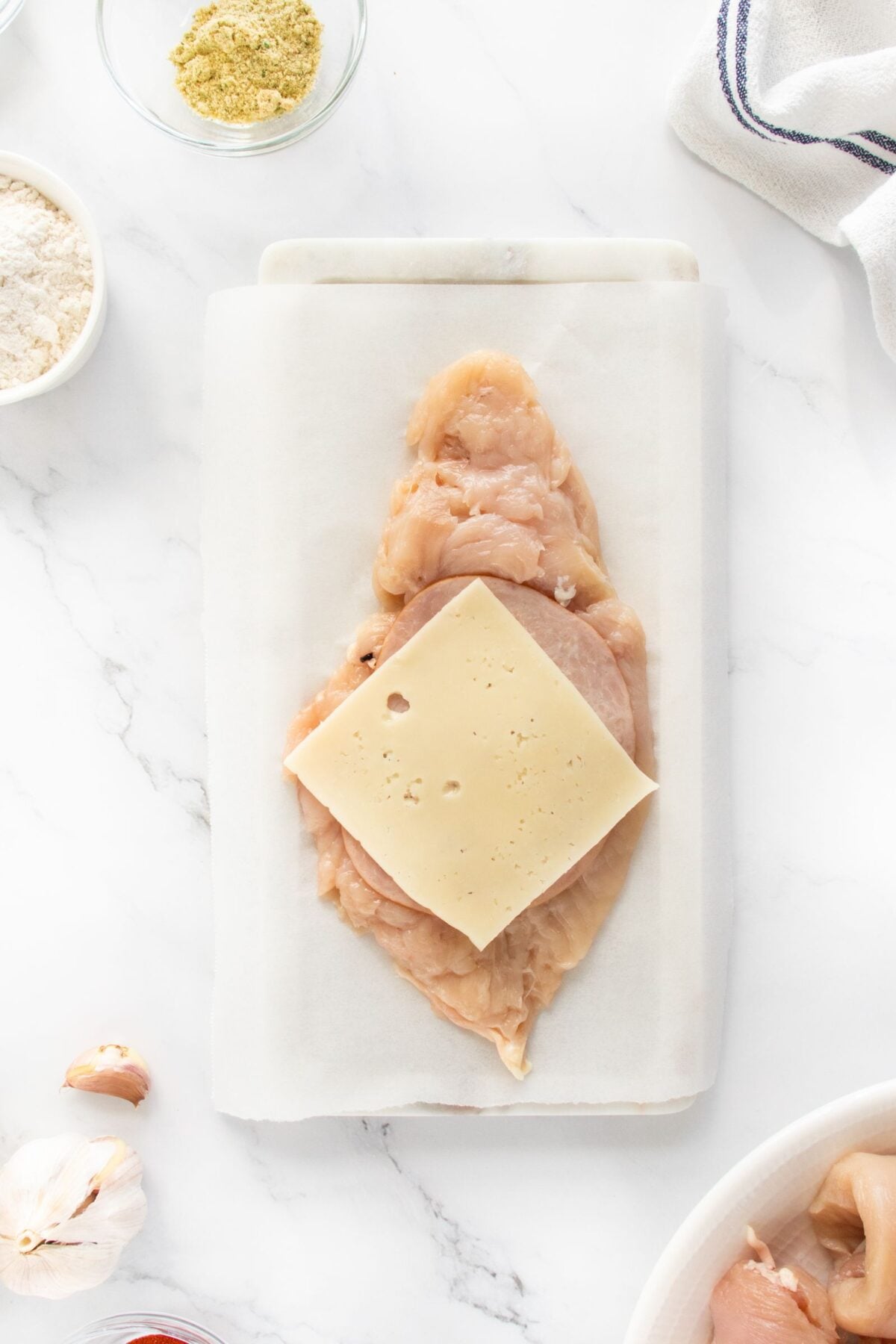 Raw chicken breast is laid open on a parchment-lined board with a slice of Swiss cheese on top. Surrounding ingredients, such as flour, spices, and garlic, are visible on a white marble surface.