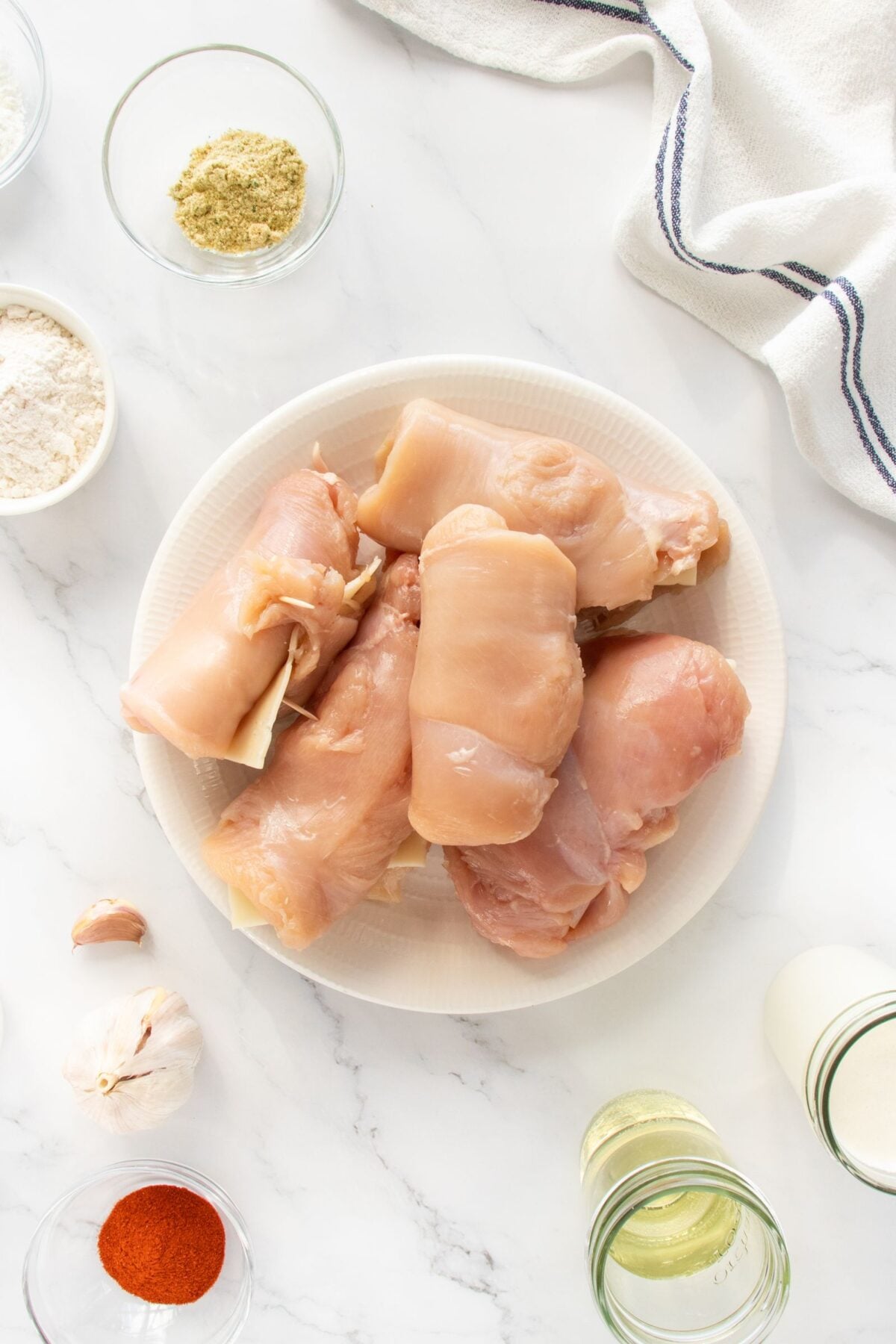 Raw chicken breasts on a white plate surrounded by small bowls of flour, spices, garlic, oil, and milk on a white marble surface, with a white kitchen towel in the corner.
