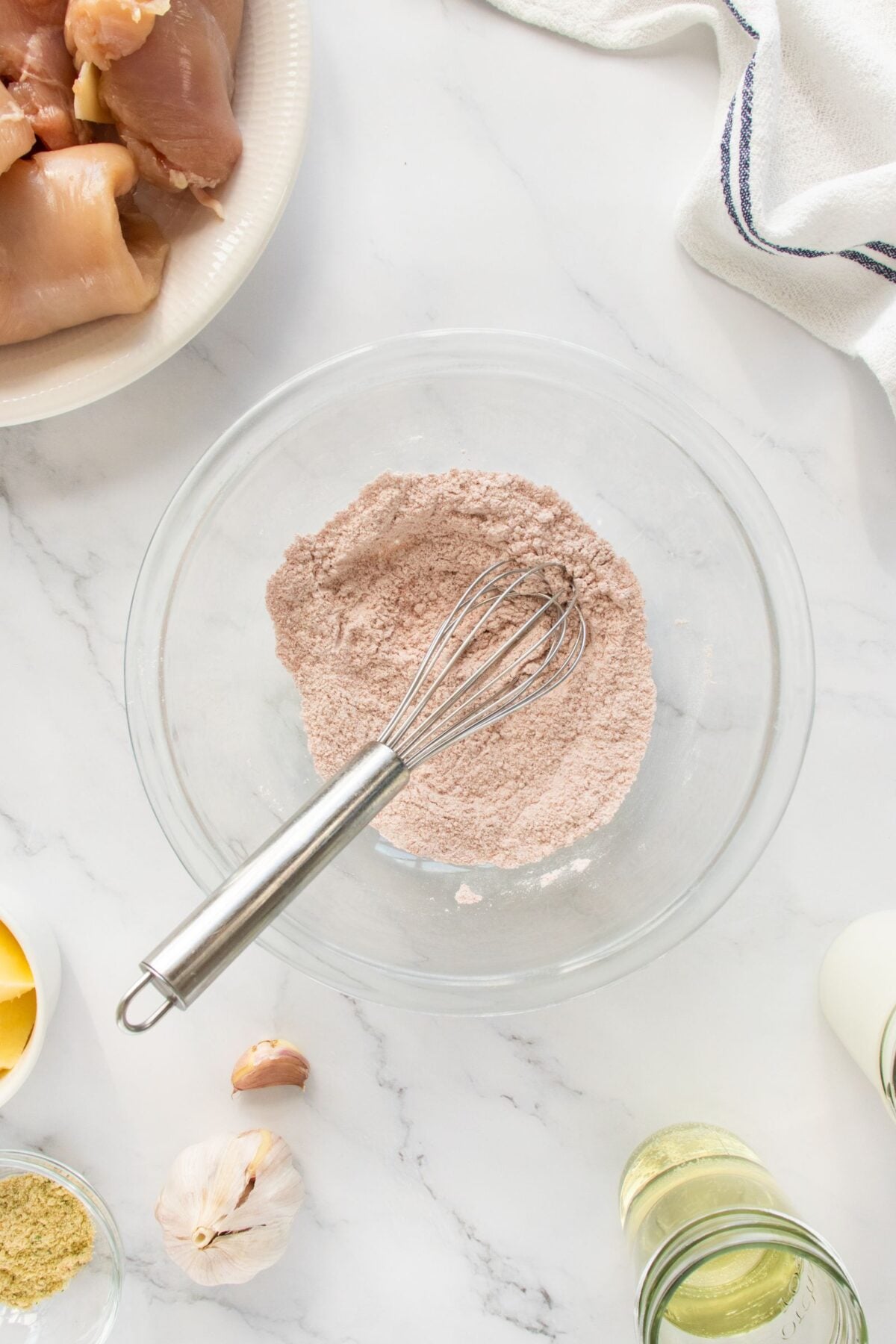 A metal whisk rests in a glass bowl filled with a reddish-brown flour mixture on a white marble surface, surrounded by raw chicken, garlic, oil, seasonings, and a white towel.