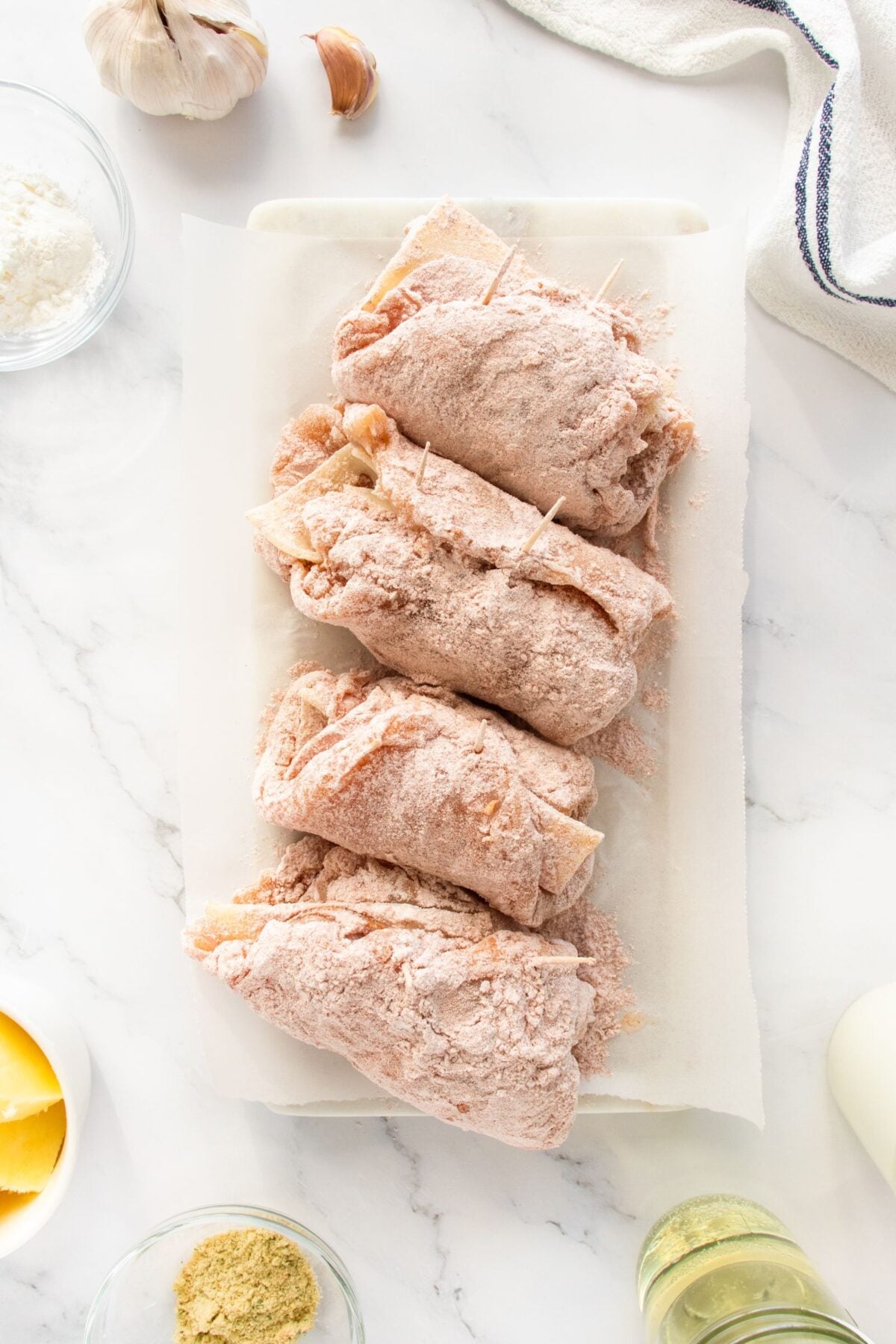 Four pieces of uncooked, breaded meat rolls secured with toothpicks are arranged on a white tray. Ingredients such as flour, garlic, broth powder, and oil are nearby on a marble countertop.