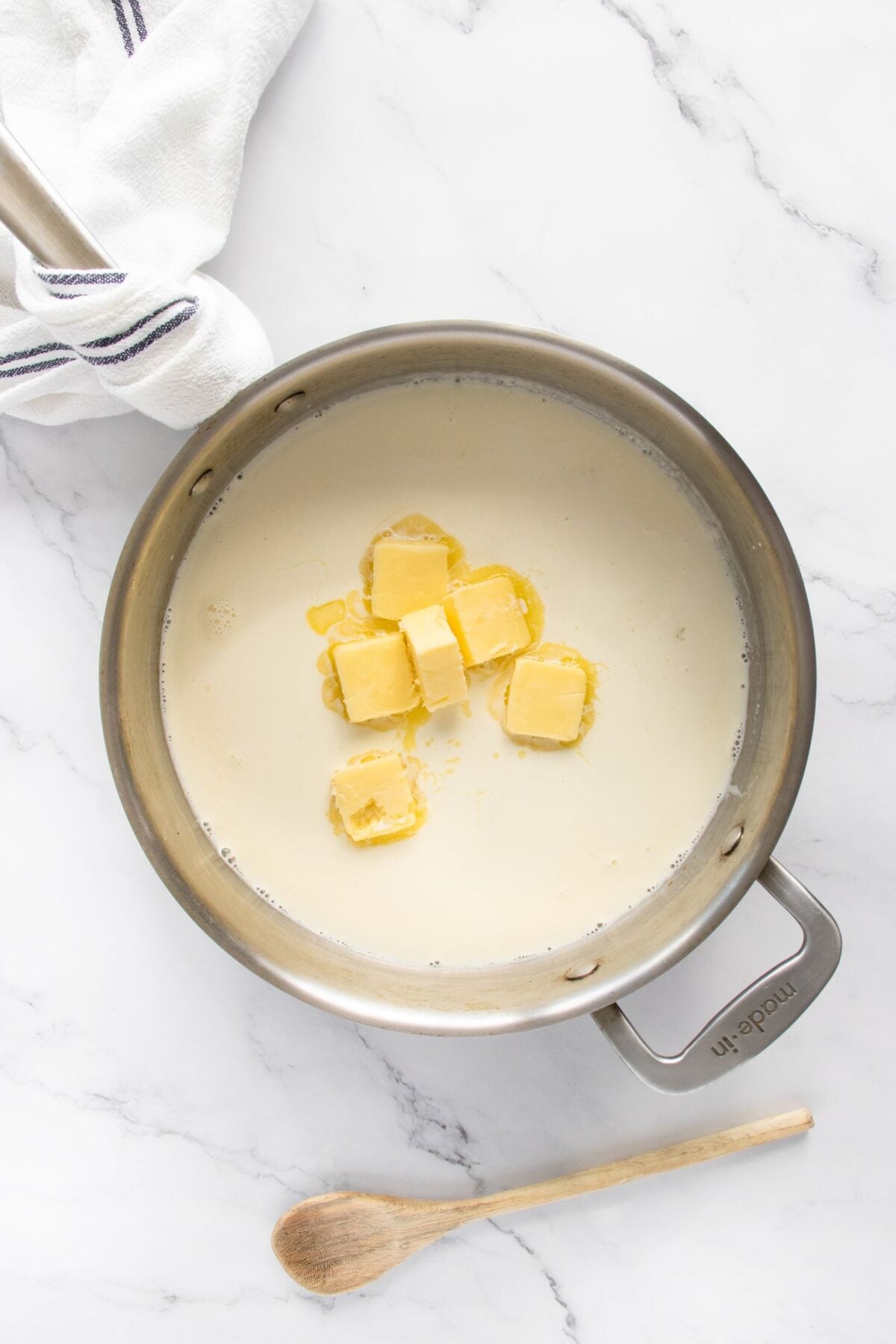 A saucepan filled with cream and several slices of butter sits on a marble countertop. A wooden spoon and a striped kitchen towel are nearby.