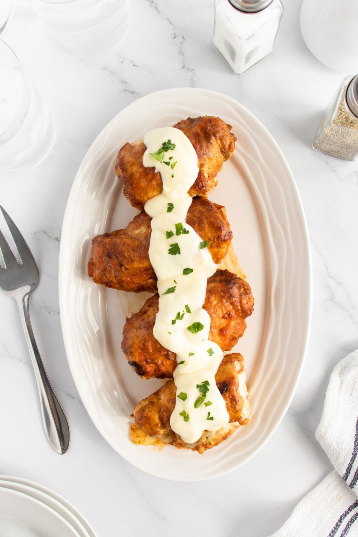Four pieces of fried chicken topped with creamy white sauce and garnished with chopped herbs on an oval white plate, set on a white marble surface with a fork, napkin, and seasonings nearby.