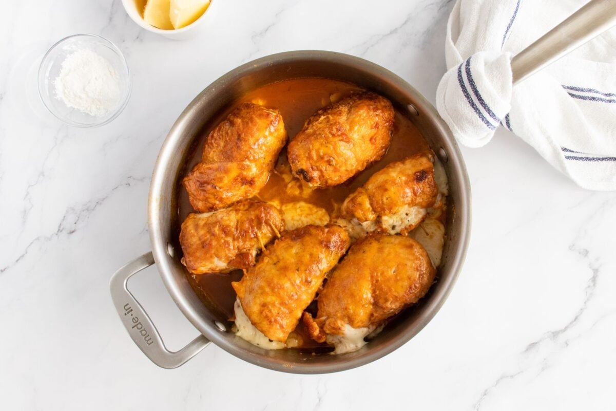 A skillet filled with golden, cooked chicken breasts in sauce sits on a marble countertop, with a bowl of flour, a bowl of butter, and a white towel with blue stripes nearby.