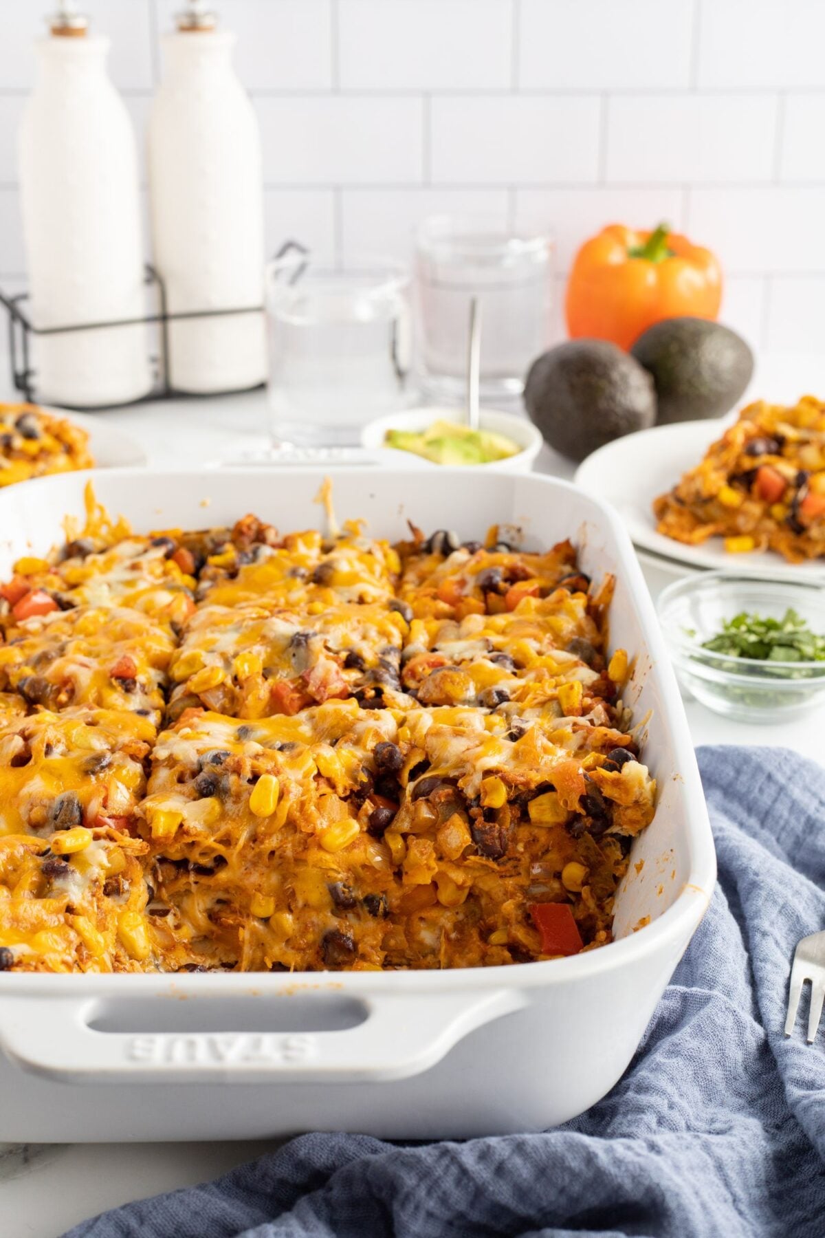 A white baking dish filled with a cheesy Mexican casserole topped with melted cheese, beans, corn, and vegetables. Slices are served on plates nearby; avocado, seasoning jars, and a tomato are in the background.
