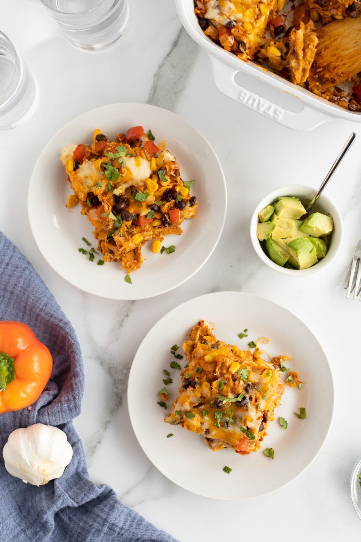 Two white plates with servings of cheesy casserole topped with chopped herbs sit on a white surface. Nearby are a bowl of diced avocado, a yellow bell pepper, a garlic bulb, and a blue napkin. A casserole dish is partially visible.