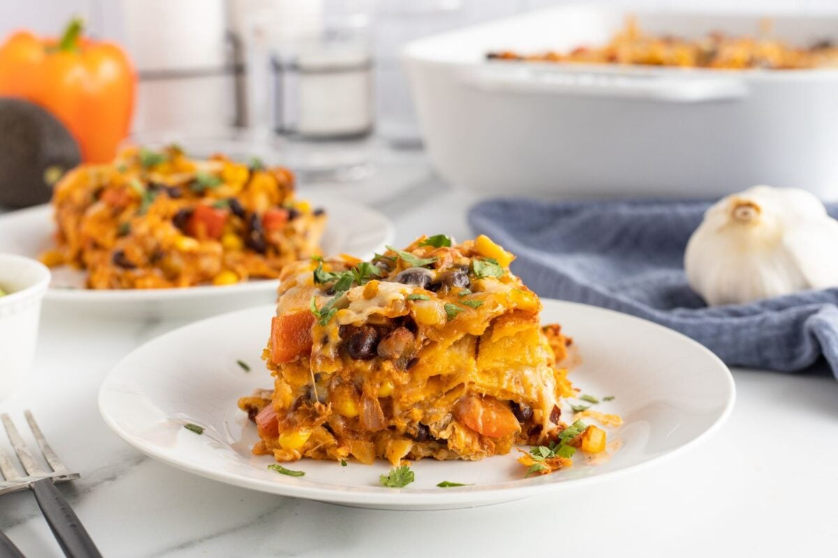 A slice of cheesy vegetable casserole with beans, corn, and tomatoes sits on a white plate, garnished with fresh herbs. A baking dish with more casserole is in the background, along with a garlic bulb and an orange bell pepper.