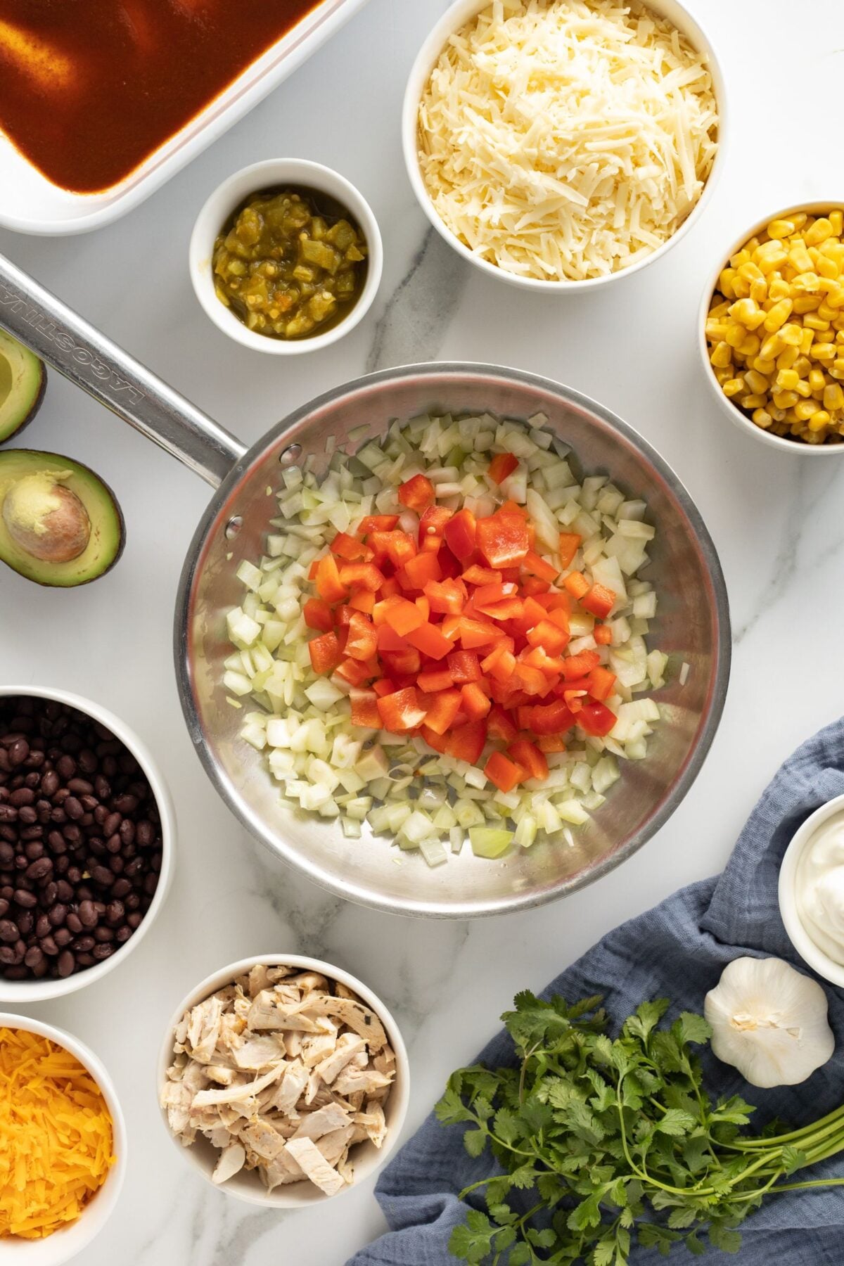 A skillet with chopped onions and tomatoes is surrounded by bowls of shredded cheese, corn, black beans, green chiles, cooked chicken, avocado, cilantro, and sour cream on a white countertop.
