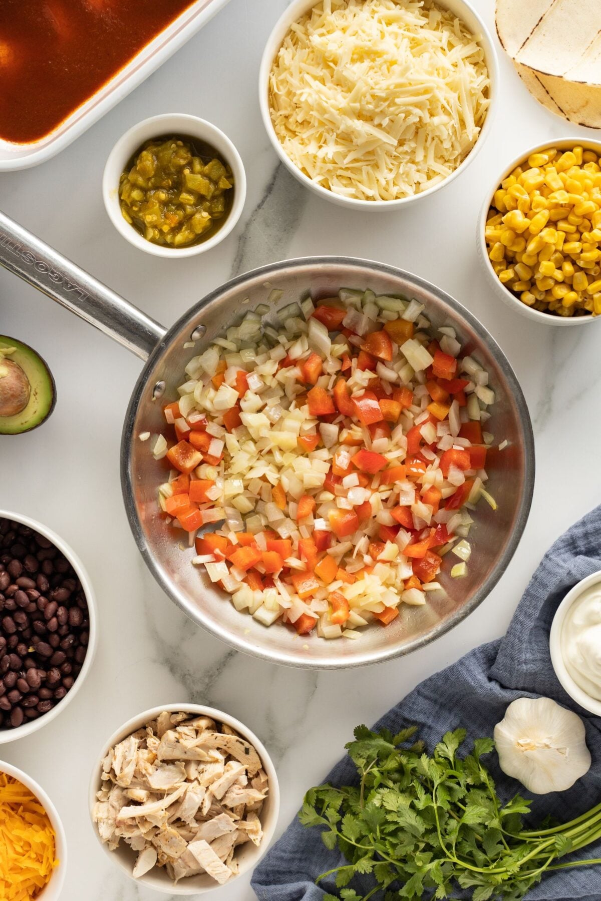 A skillet with chopped onions and tomatoes is at the center, surrounded by bowls of grated cheese, corn, black beans, sour cream, diced chicken, green chilies, and other taco ingredients on a white surface.