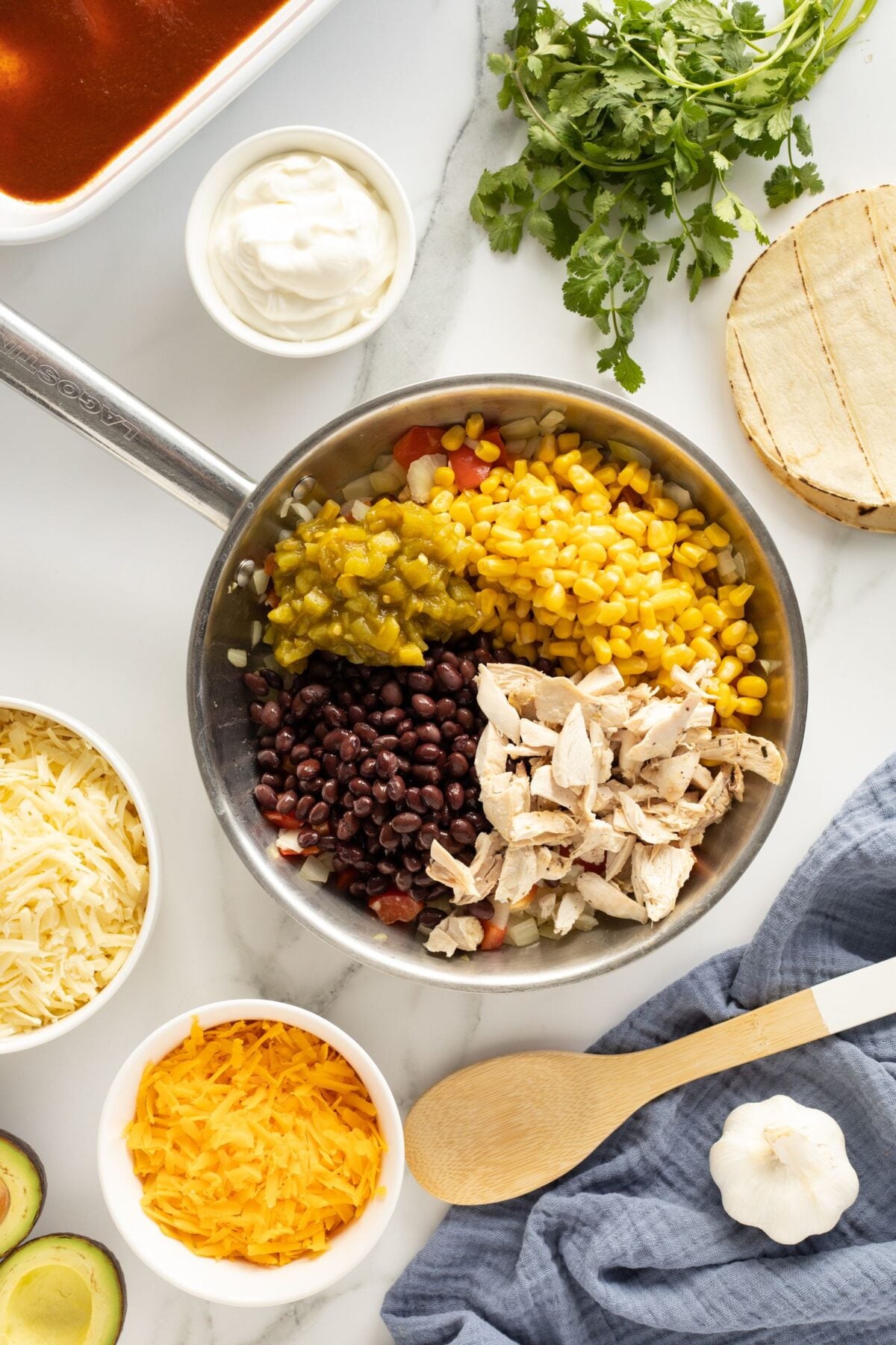 A pan filled with corn, black beans, green chilies, and shredded chicken sits on a marble counter surrounded by shredded cheese, sour cream, cilantro, tortillas, avocado, garlic, and a wooden spoon.