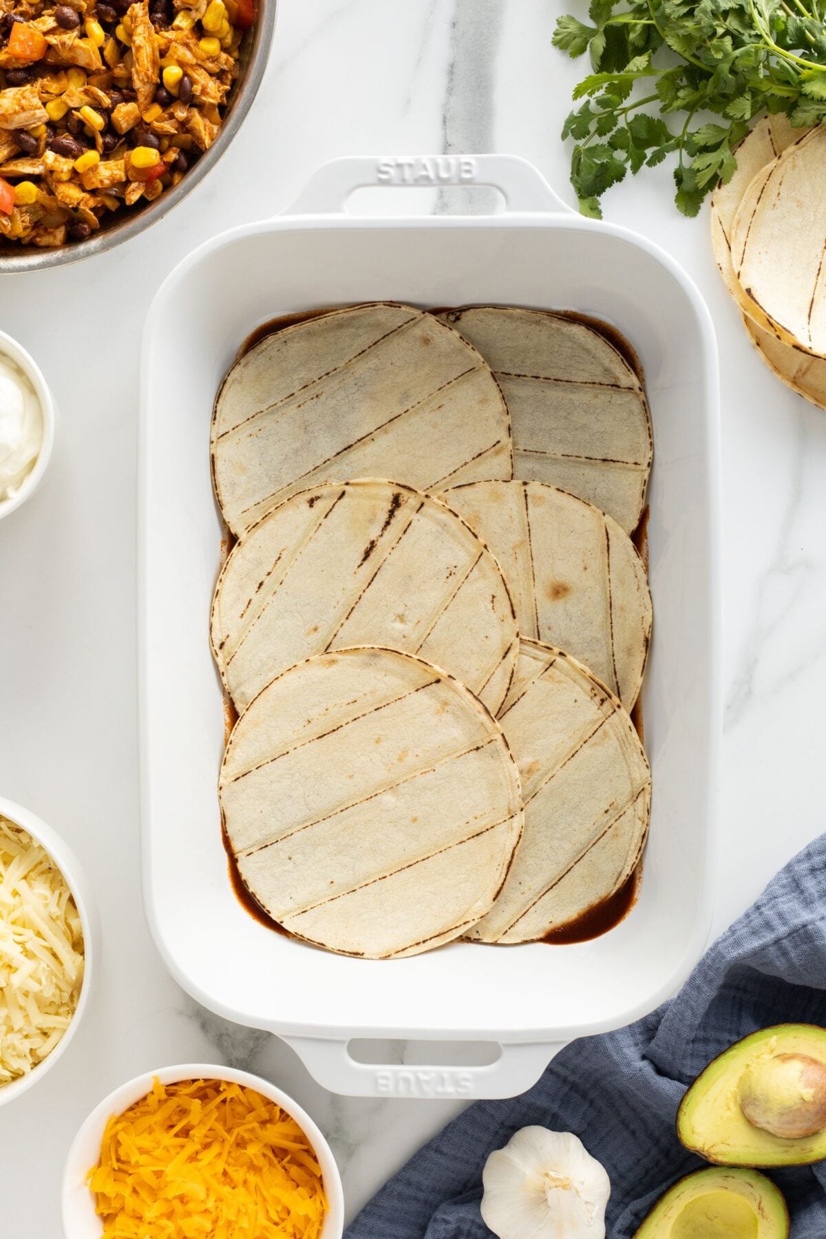 A white baking dish with six corn tortillas layered over red sauce. Surrounding the dish are bowls of shredded cheese, cooked filling, sliced onions, tortillas, fresh cilantro, avocado, and a garlic bulb on a marble counter.