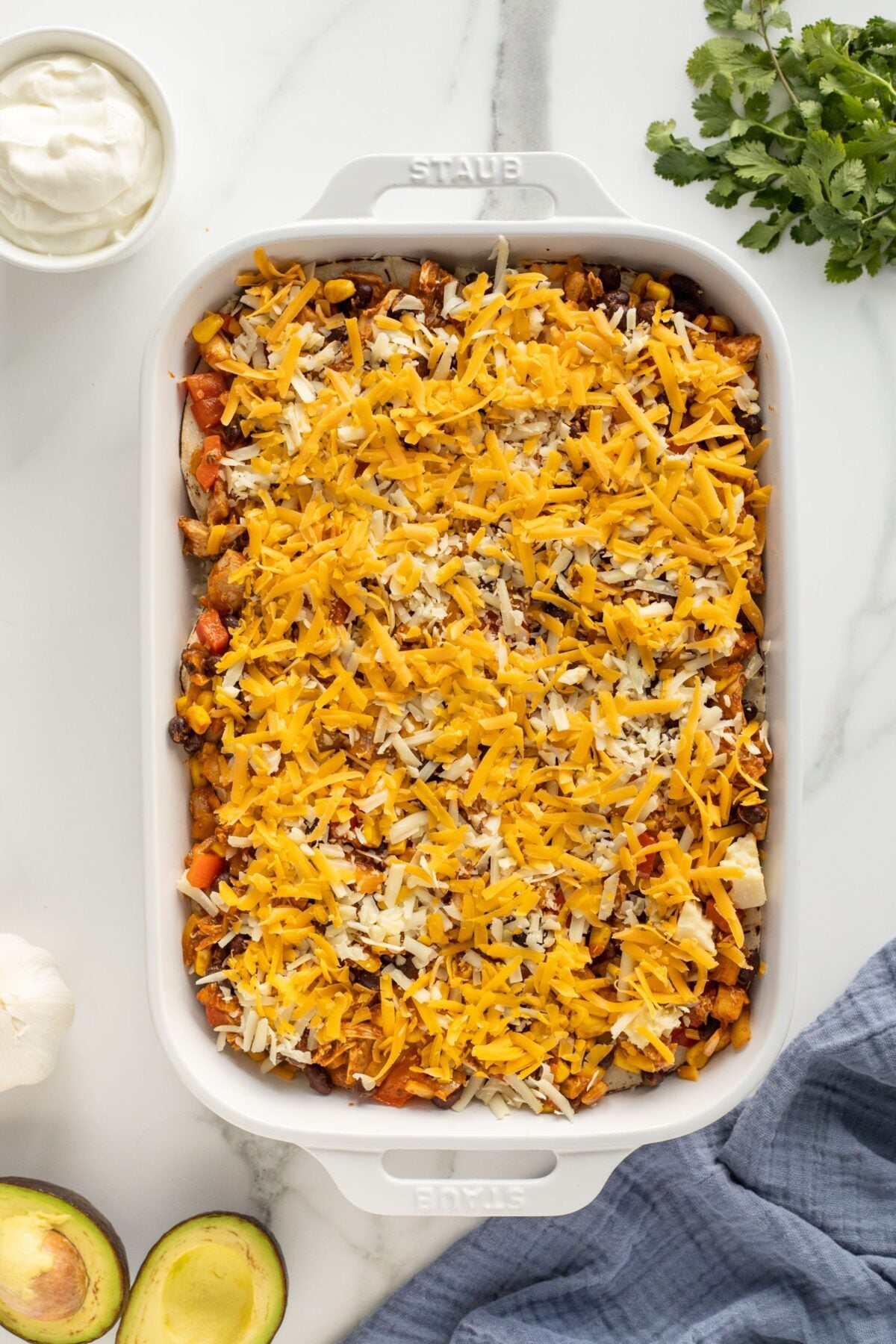 A white baking dish filled with a layered casserole topped with shredded cheddar and mozzarella cheeses sits on a marble surface surrounded by avocado halves, cilantro, sour cream, garlic, and a blue cloth.