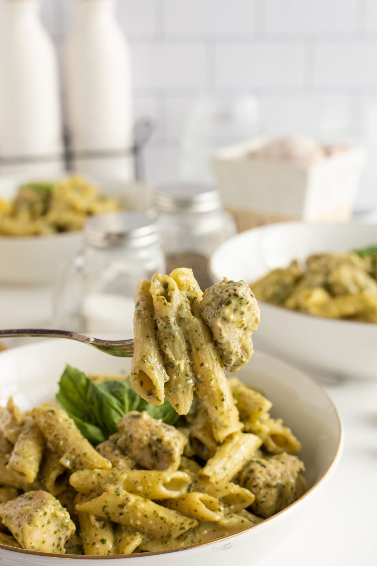 A close-up of a fork holding penne pasta with creamy pesto sauce and pieces of chicken, above a bowl filled with more pasta. Bowls and condiments are blurred in the background.