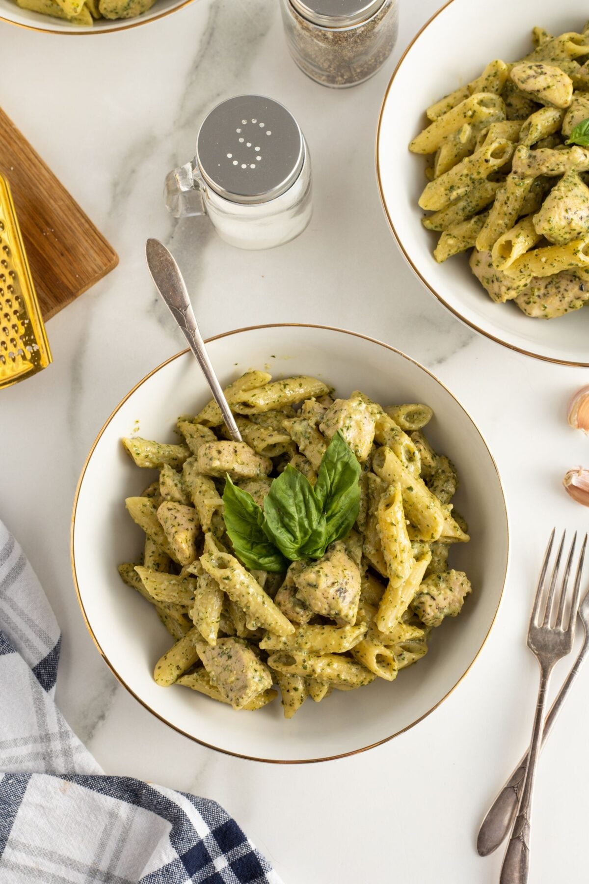 A bowl of penne pasta with pesto sauce and pieces of chicken, garnished with fresh basil. Other bowls of pasta, a salt shaker, a cheese grater, and utensils are visible on a white marble surface.