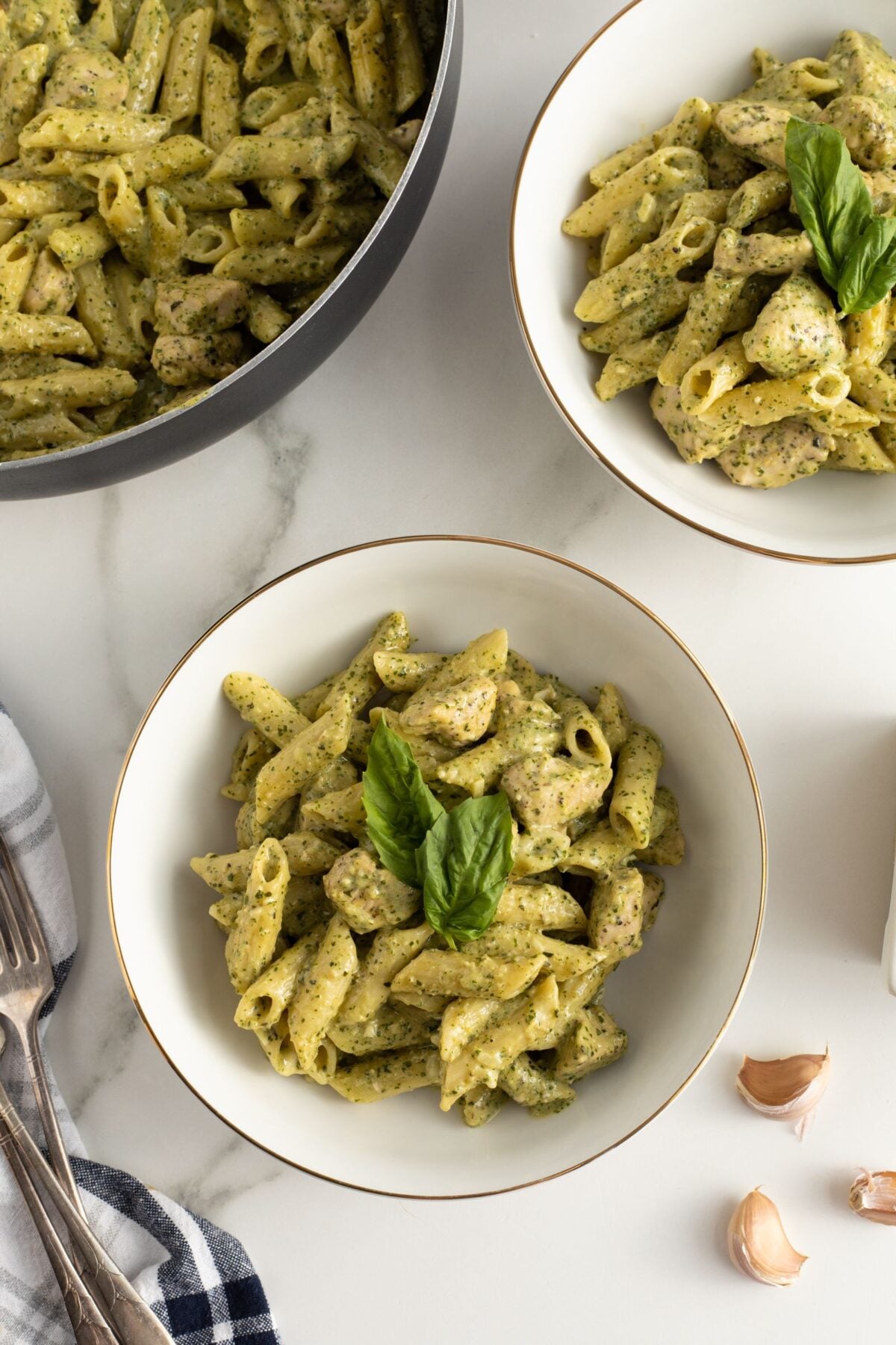 Three bowls of penne pasta with pesto sauce and pieces of chicken, garnished with fresh basil leaves, are arranged on a white surface next to a pan of pasta, forks, a cloth napkin, and garlic cloves.