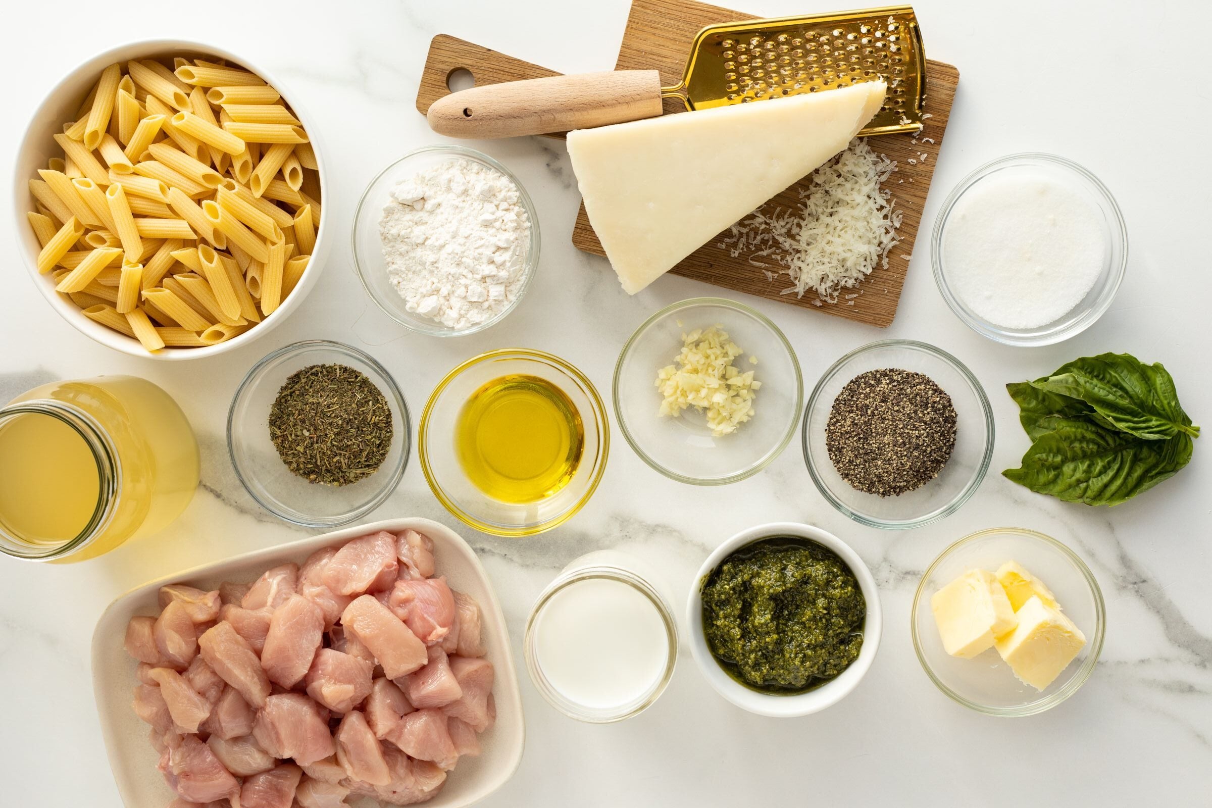 Overhead view of uncooked chicken pieces, dry penne pasta, grated cheese, basil, seasonings, butter, olive oil, flour, pesto, milk, broth, minced garlic, and sugar arranged in bowls on a white surface.
