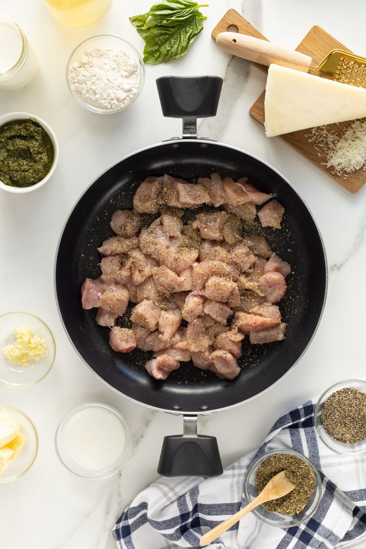 Overhead view of a pan with seasoned raw chicken pieces, surrounded by bowls of pesto, cream, minced garlic, butter, flour, and herbs, with a block of Parmesan and basil on a white marble surface.