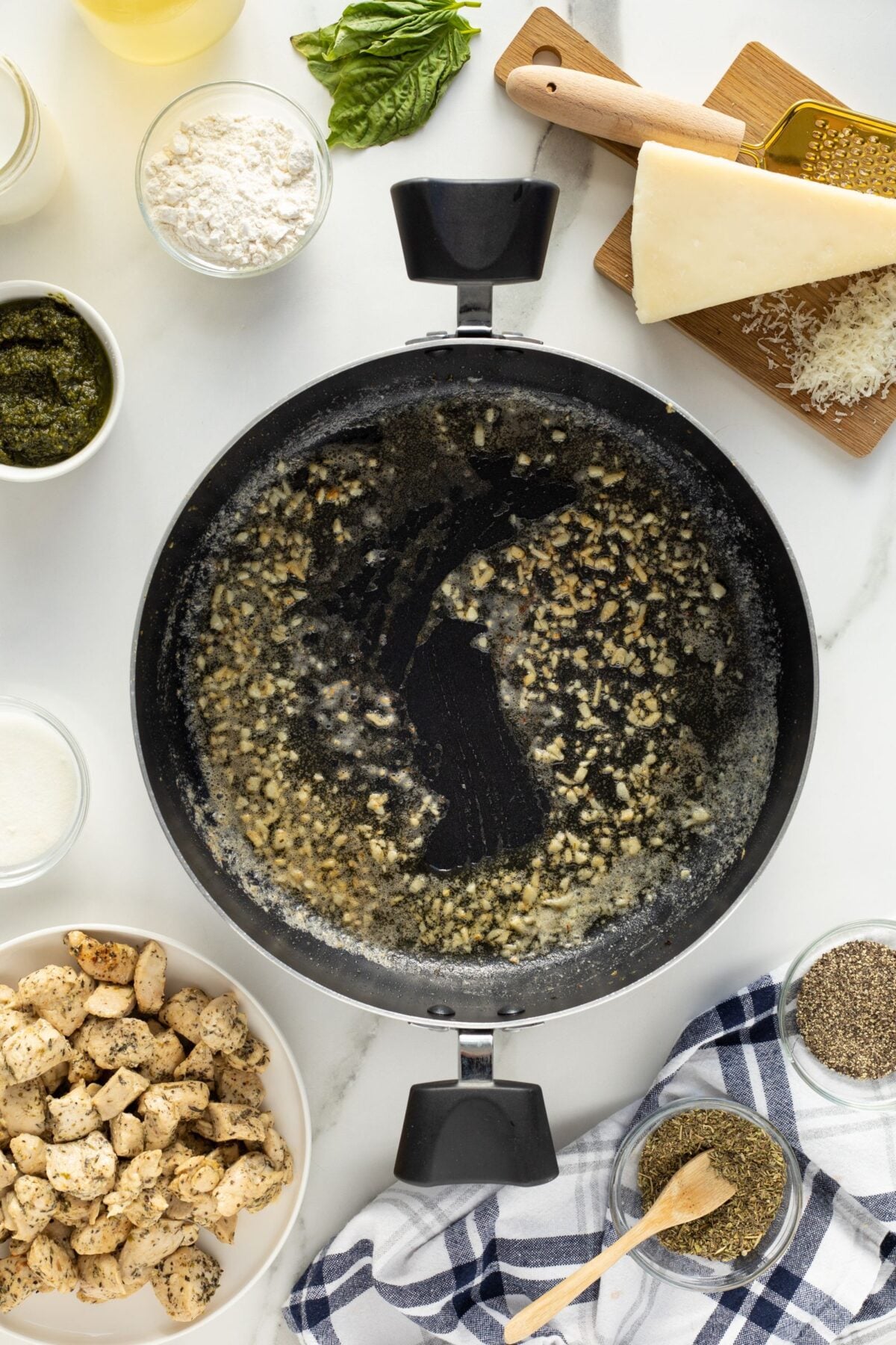 A skillet with sizzling minced garlic in melted butter, surrounded by ingredients for cooking, including shredded cheese, cream, flour, pesto, cooked chicken, spices, and fresh basil on a white countertop.