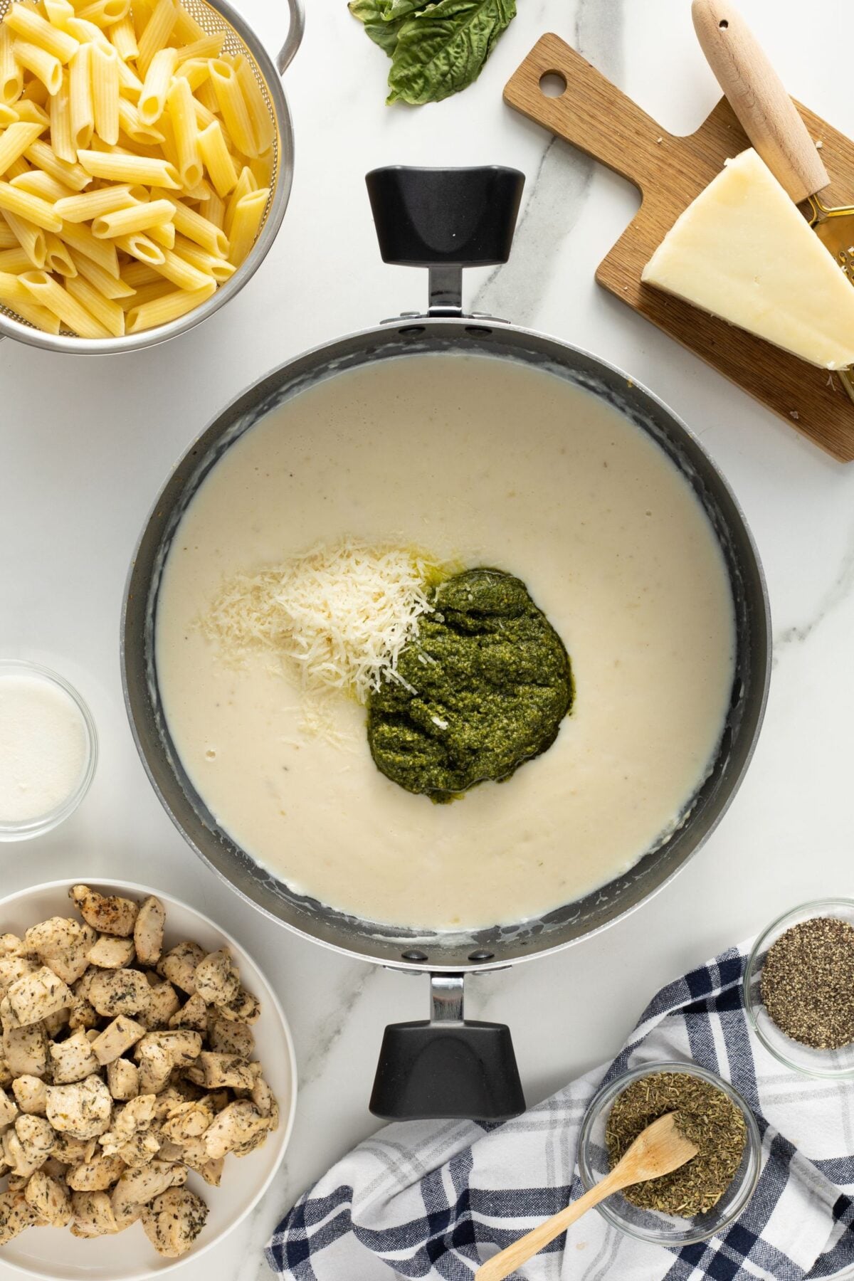A pot of creamy white sauce with shredded cheese and pesto on top, surrounded by cooked penne pasta, cooked chicken, grated cheese, spices, a milk jug, basil leaves, and a blue plaid towel on a white surface.
