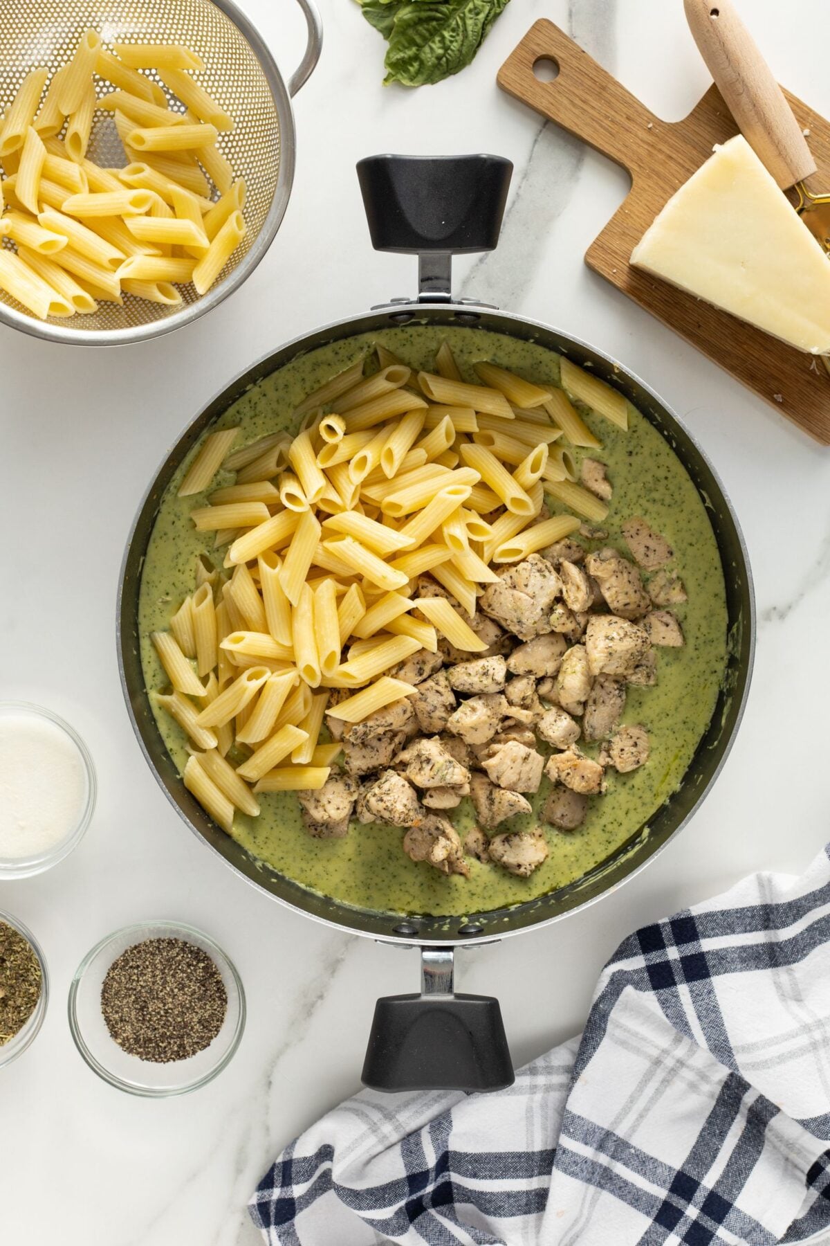 A skillet containing cooked penne pasta and chunks of seasoned chicken atop creamy green pesto sauce, surrounded by a bowl of penne, grated cheese, herbs, and a checkered kitchen towel on a white countertop.