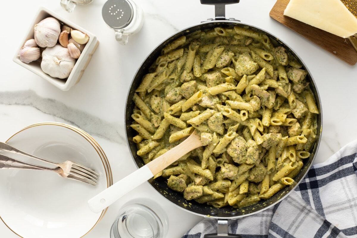 A skillet filled with penne pasta and chicken in a green pesto sauce sits on a marble counter. Nearby are garlic bulbs, plates with cutlery, a wedge of cheese, a glass of water, and a checkered napkin.