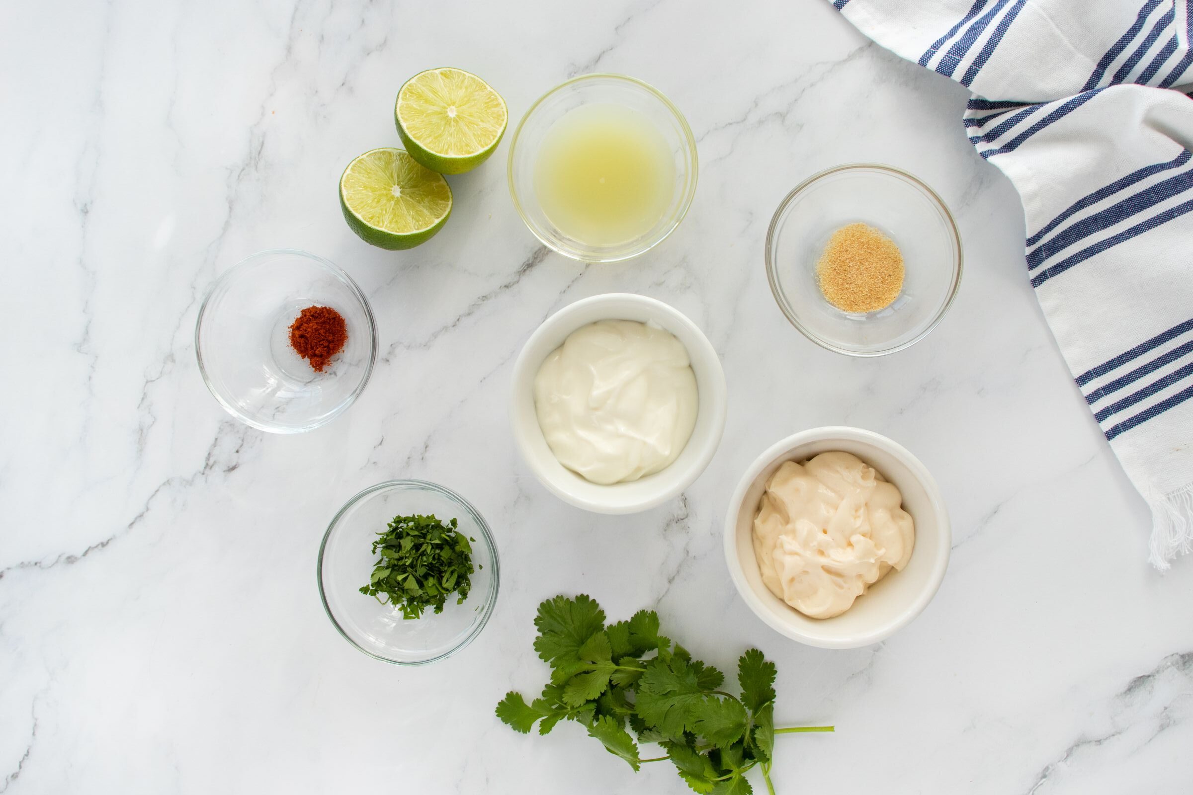 A marble countertop with small bowls containing chopped herbs, red spice, lime halves, lime juice, mayonnaise, sour cream, and garlic powder, alongside fresh cilantro and a striped kitchen towel.