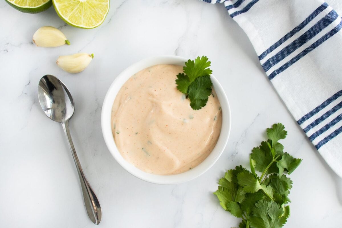 A bowl of creamy sauce garnished with cilantro sits on a marble surface. Nearby are fresh cilantro, garlic cloves, lime halves, a spoon, and a white napkin with blue stripes.