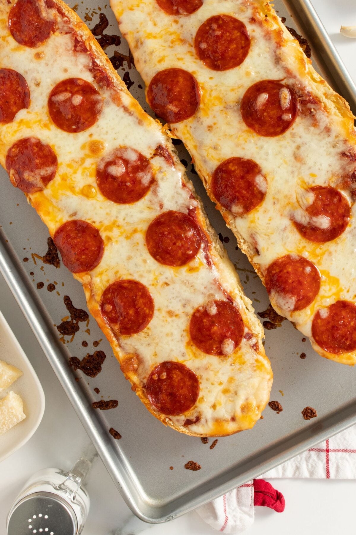 Two loaves of French bread pizza topped with melted cheese and pepperoni slices are on a baking sheet. Grated cheese, a pizza cutter, and a red-striped napkin are nearby.