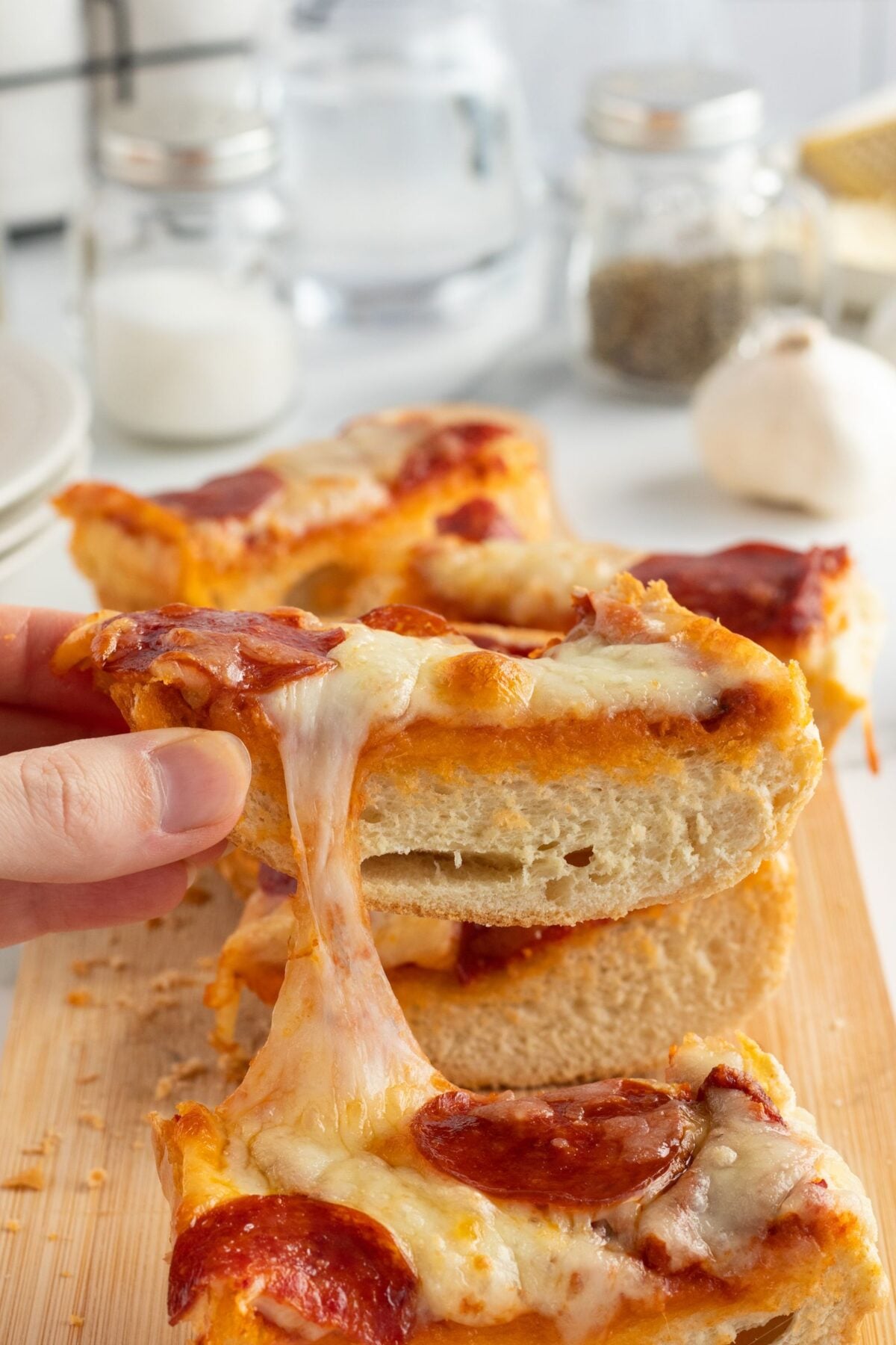 A hand pulls a cheesy slice of pepperoni pizza from a stack on a wooden board, with melted cheese stretching between slices. Seasoning jars, garlic, and a glass of water are visible in the background.