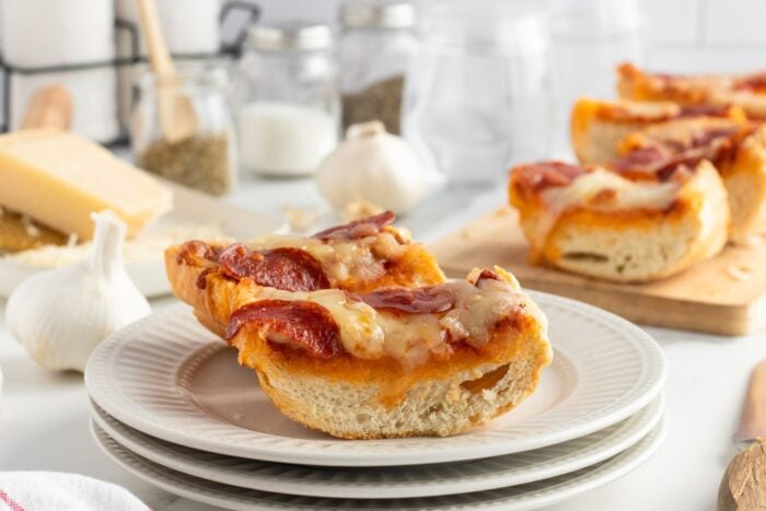 Slices of French bread pizza topped with melted cheese and pepperoni are stacked on white plates, with garlic, cheese, and seasoning jars in the background on a white kitchen counter.