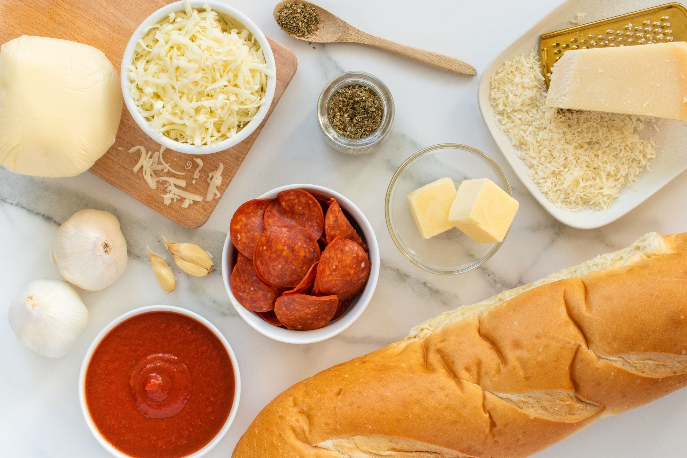 A baguette, pepperoni slices, tomato sauce, shredded cheese, garlic, grated parmesan, Italian seasoning, and a block of mozzarella on a marble surface, ready for making pizza.
