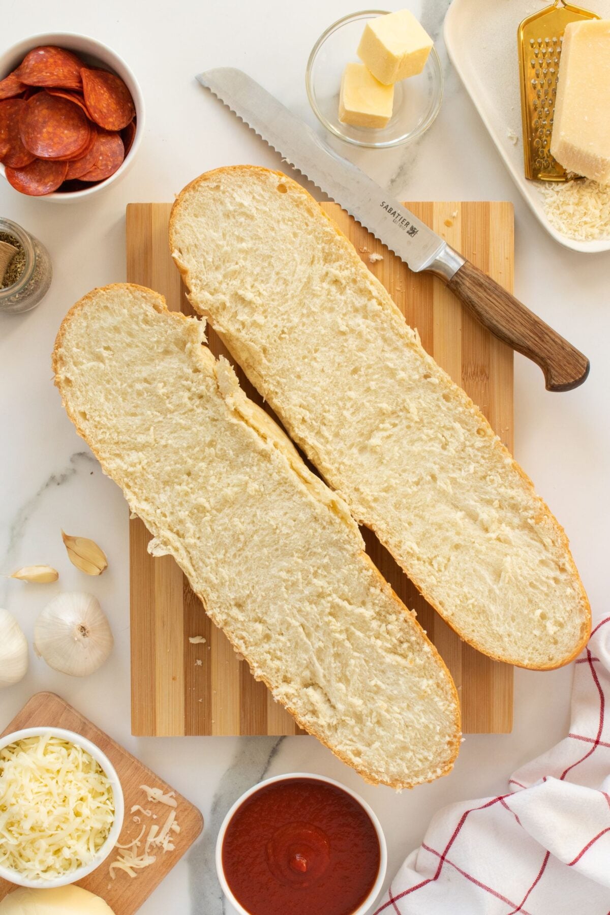 A sliced loaf of bread on a cutting board is surrounded by pizza ingredients: pepperoni, butter, cheese, garlic, tomato sauce, grated parmesan, and a knife.