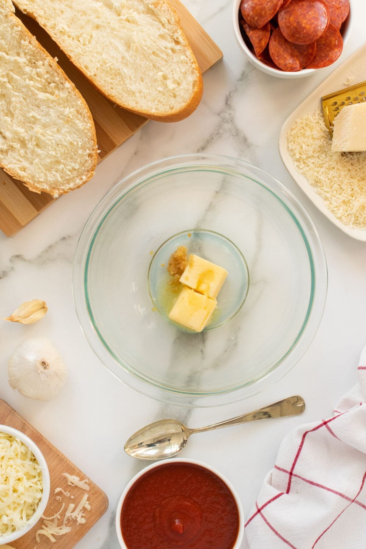 A glass bowl with butter and minced garlic is surrounded by sliced bread, shredded cheese, sliced pepperoni, grated parmesan, marinara sauce, a garlic bulb, and a spoon on a marble countertop.