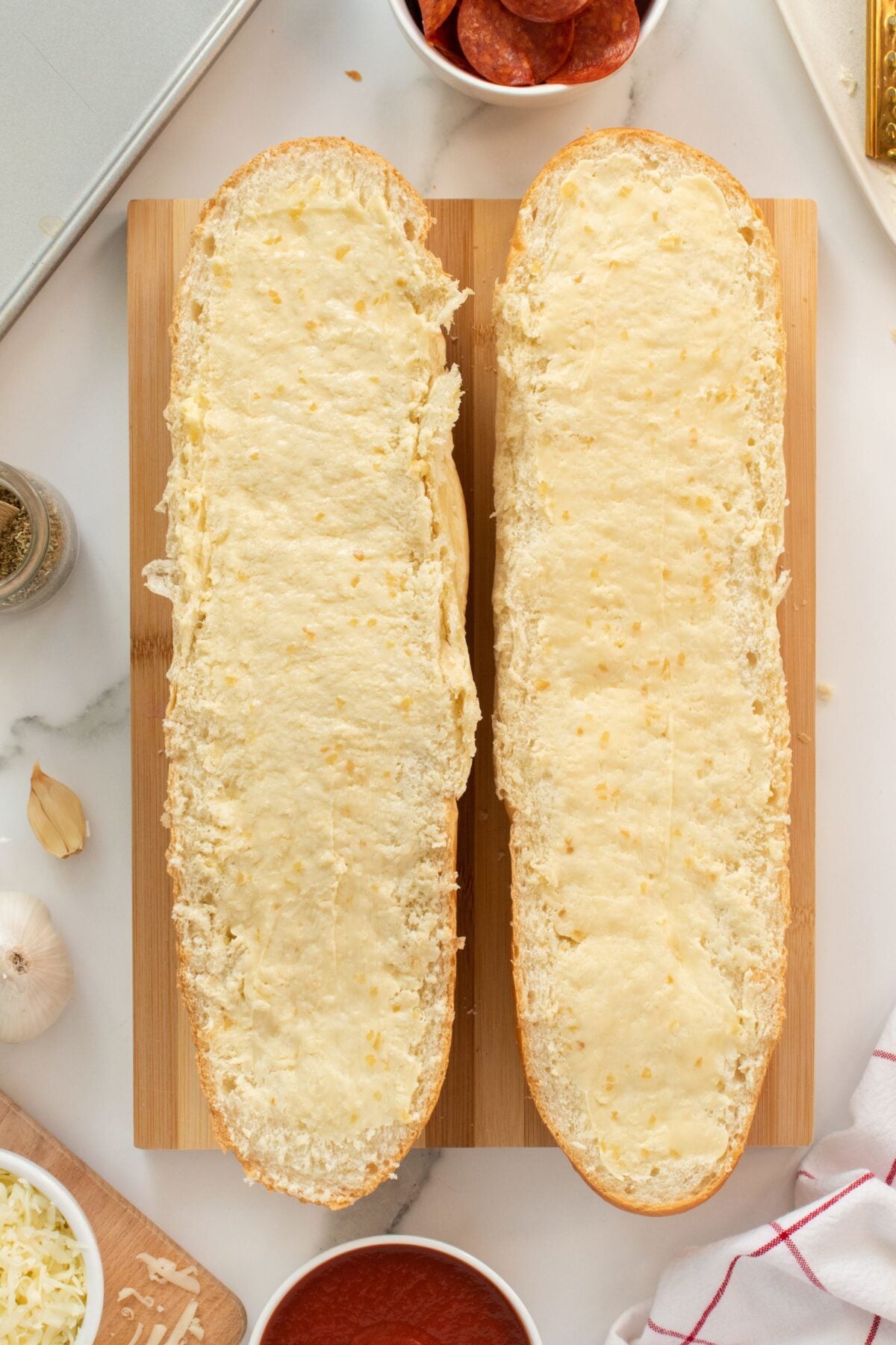 Two halves of a loaf of bread are topped with melted cheese and placed side by side on a wooden cutting board, surrounded by ingredients like garlic, pizza sauce, pepperoni, and shredded cheese.