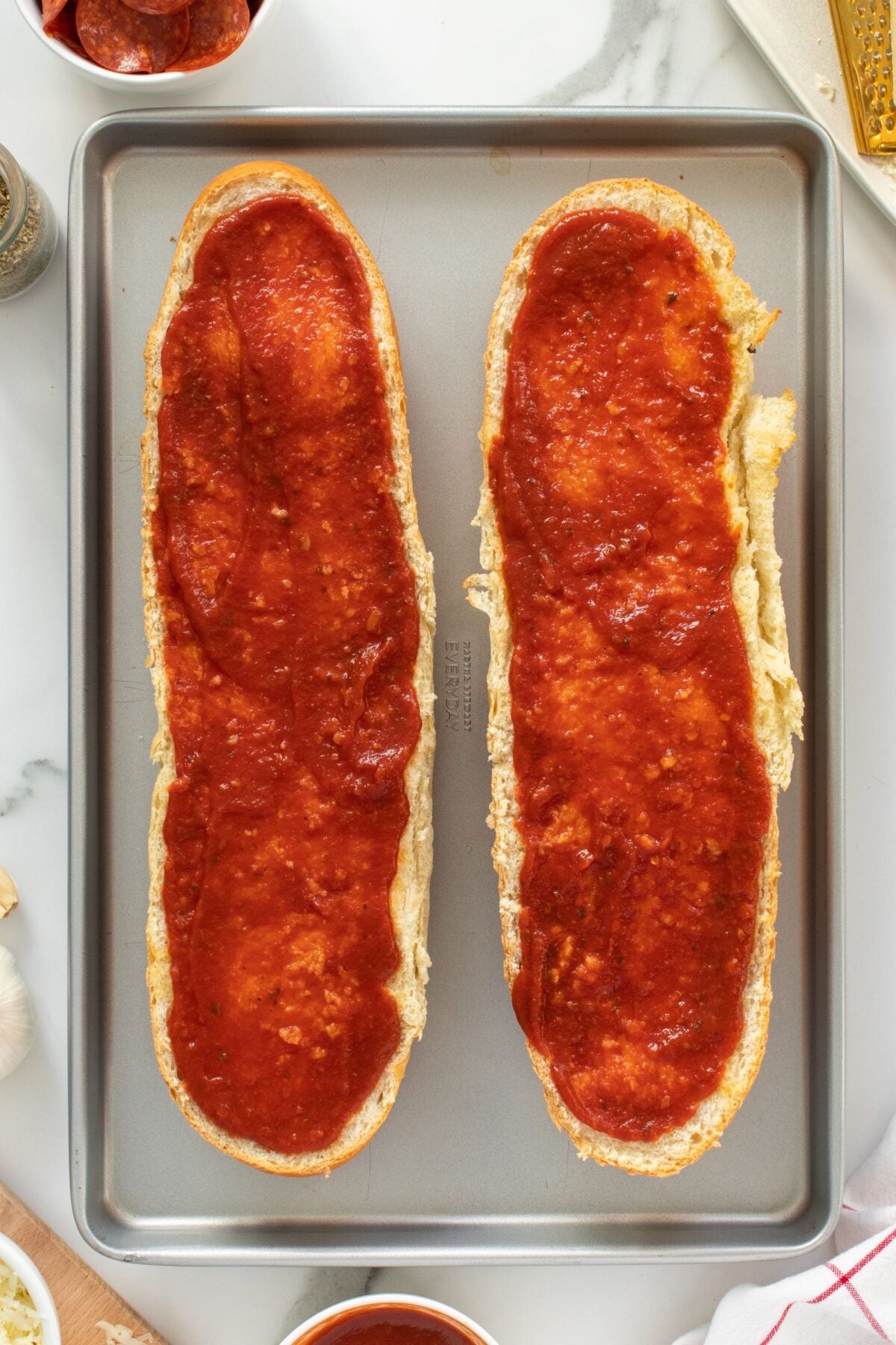 A baking sheet holds two halves of French bread, each topped with a layer of red pizza sauce, ready for additional toppings. Ingredients and utensils are partially visible around the tray.