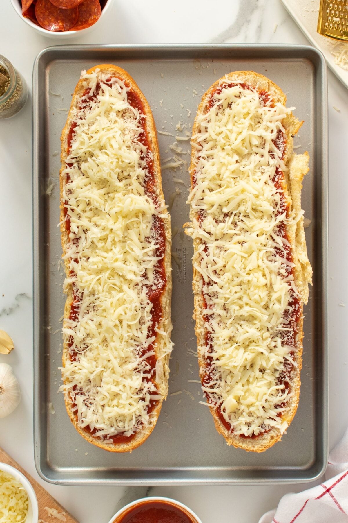 Two halves of a French bread loaf topped with pizza sauce and shredded mozzarella cheese sit on a baking sheet, ready to be baked. Ingredients like pepperoni, garlic, and grated cheese are visible nearby.