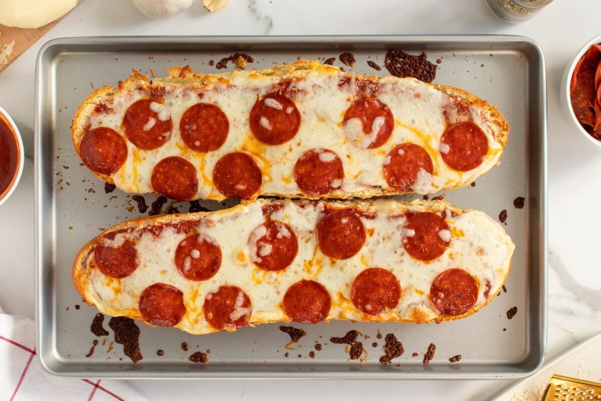 Two halves of French bread topped with melted cheese and pepperoni slices, baked on a metal tray, with scattered sauce and ingredient bowls visible around the edges.