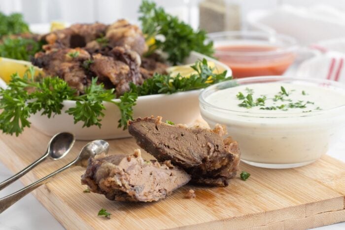 Close-up of crispy fried pieces of meat on a wooden board, with parsley garnish, a bowl of creamy white dipping sauce topped with herbs, and small metal spoons beside them. Blurred ketchup and more food in the background.