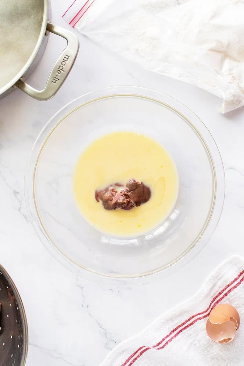 A glass mixing bowl contains melted butter and a small amount of chocolate on a white marble countertop. Nearby are a saucepan, a cracked eggshell, and a white towel with a red stripe.