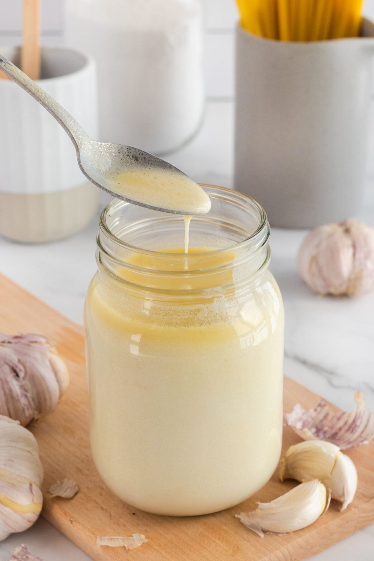 A spoon drizzles creamy white sauce into a glass jar on a wooden board, surrounded by garlic bulbs. In the background are kitchen containers and uncooked spaghetti.
