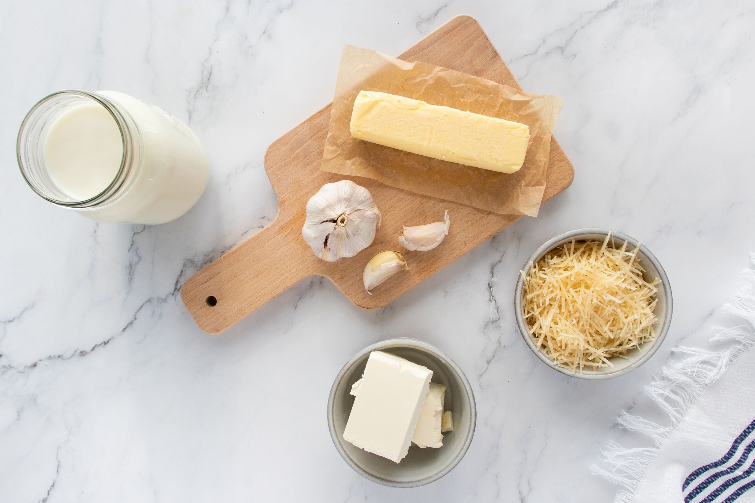 A block of butter on parchment, a head of garlic with cloves, a jar of milk, grated cheese in a bowl, and cream cheese in a bowl, all arranged on a white marble surface.