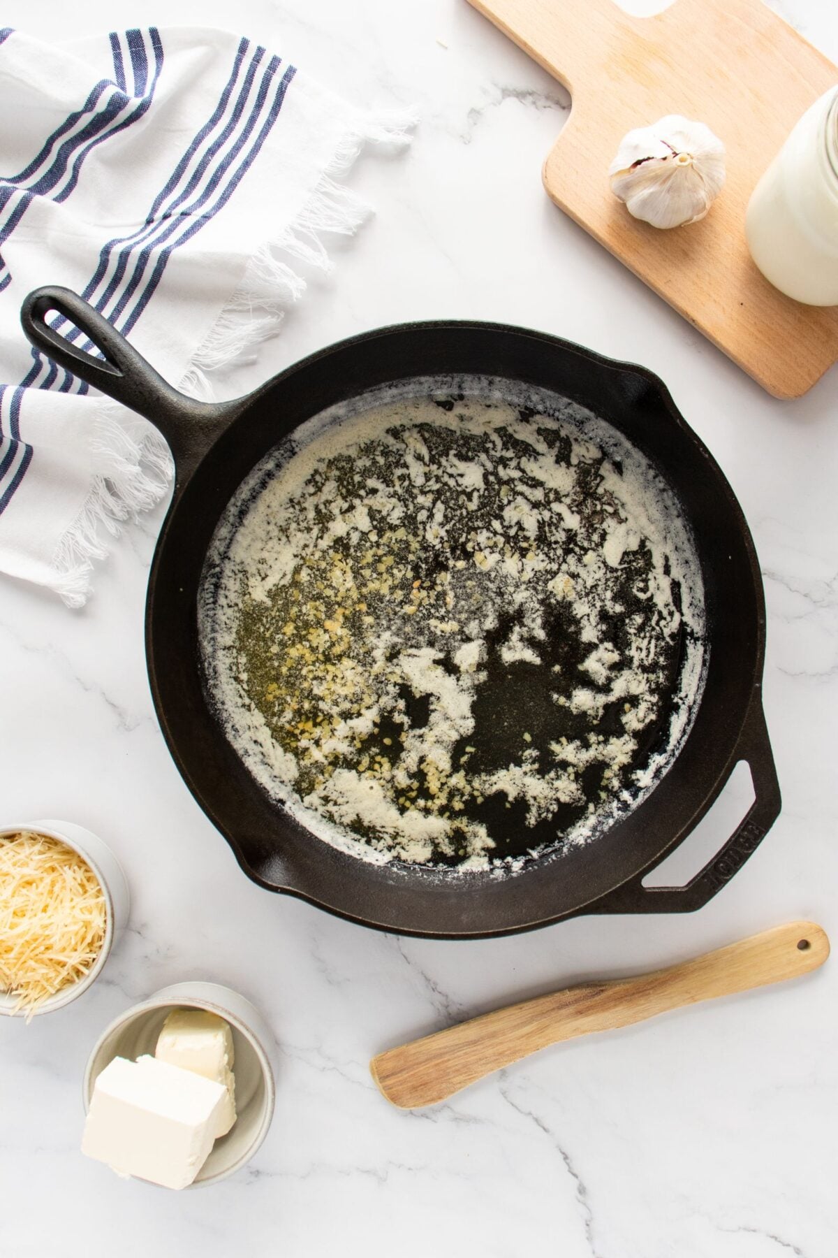 A black cast iron skillet with melted butter sits on a marble countertop, surrounded by a wooden spoon, a bowl of shredded cheese, a dish of cream cheese, a jar of milk, a garlic bulb, and a striped towel.